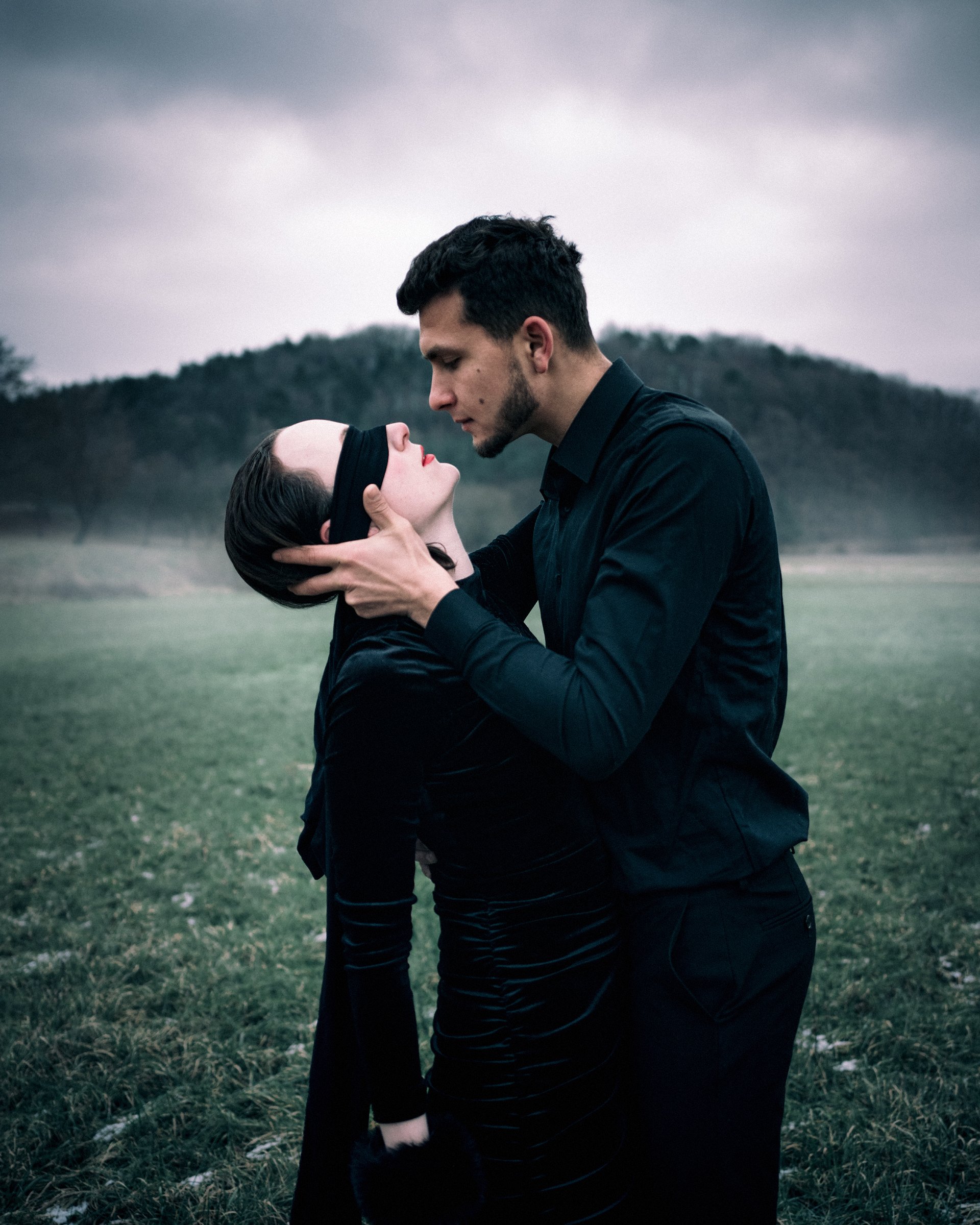 A man and a woman, both dressed in black, standing in a grassy field with a mountainous background, are engaged in a moment of intimacy. The woman is wearing a blindfold, and the man is holding her face with both hands as they lean close to each othe