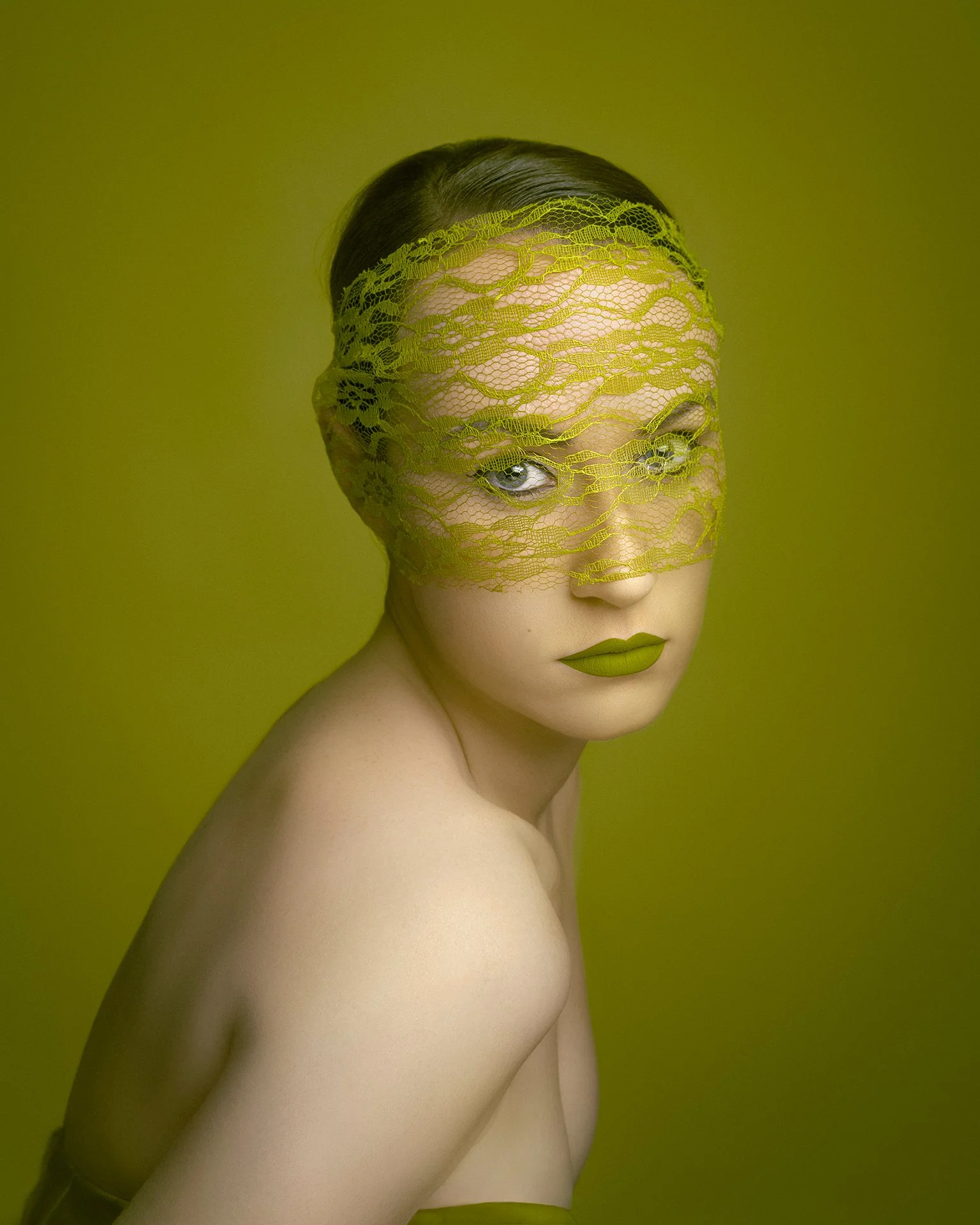 A woman with fair skin and dark hair wearing green lipstick and a green lace mask covering part of her face, against a matching green background.