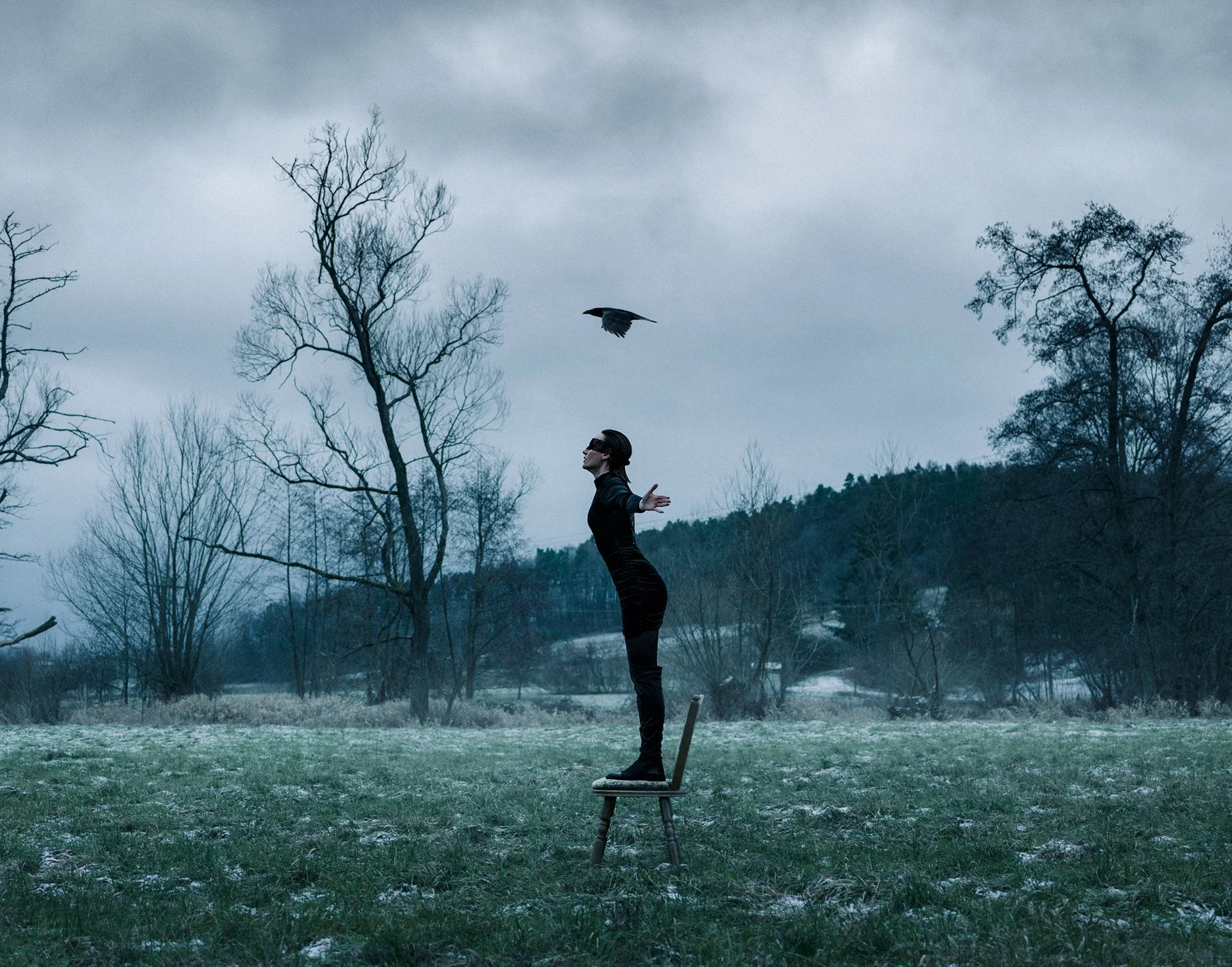 A woman standing on a chair outside in a field with leafless trees, with her arms outstretched, as a bird flies above her on a cloudy day.