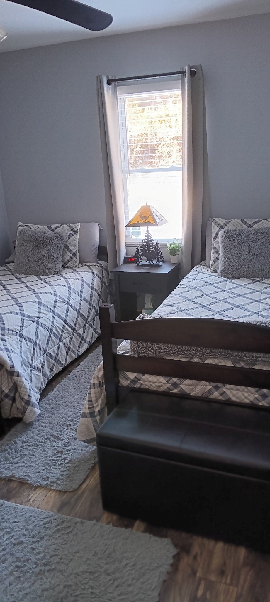 Cozy bedroom with two beds, plaid bedding, furry cushions, and a dark wooden bench. A table with a decorative lamp and a potted plant is between the beds, next to a window with curtains.