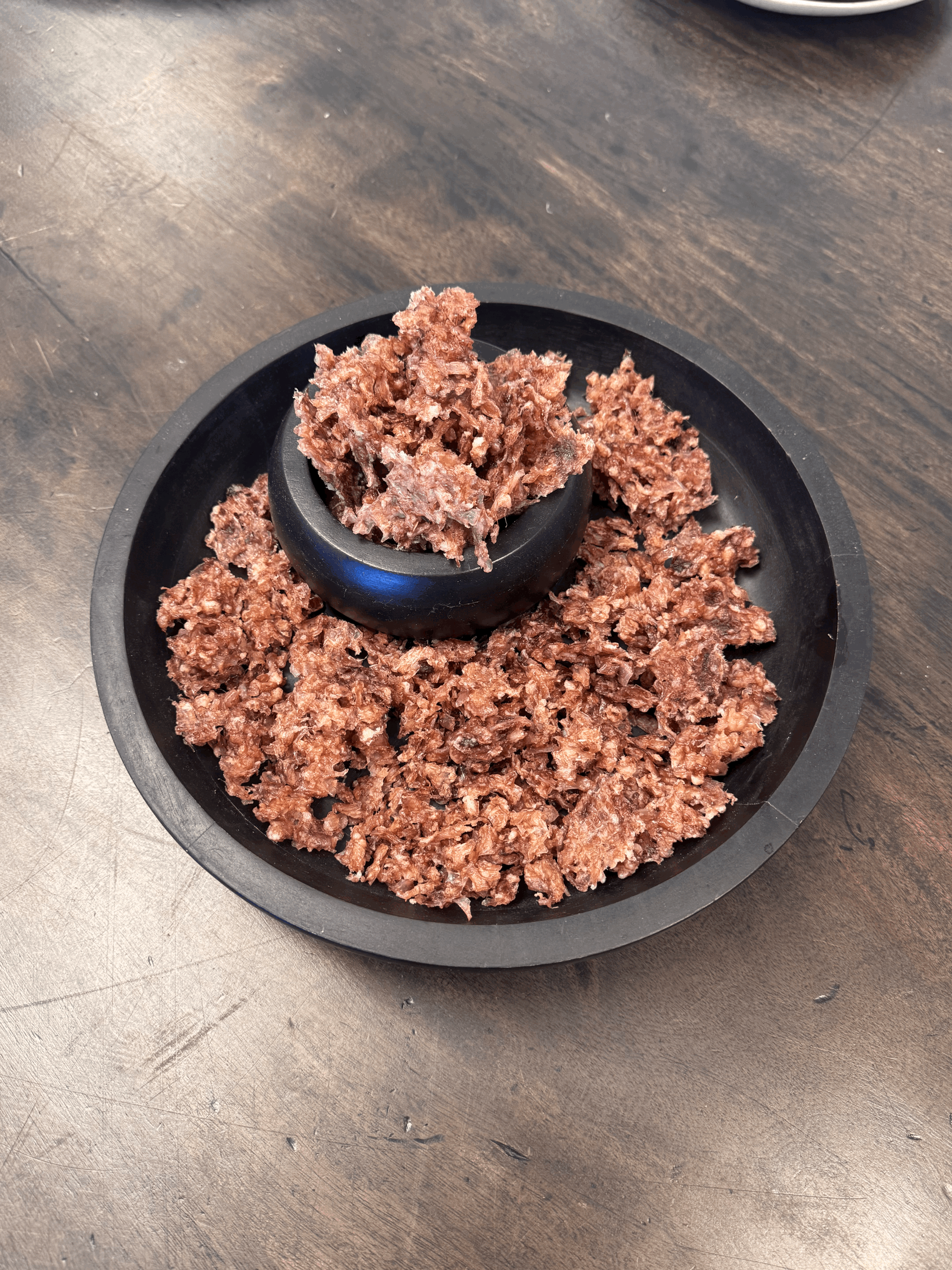 Air-Dried Lamb Trachea Crumble displayed in a small black bowl surrounded by crumbled pieces on a round black platter.