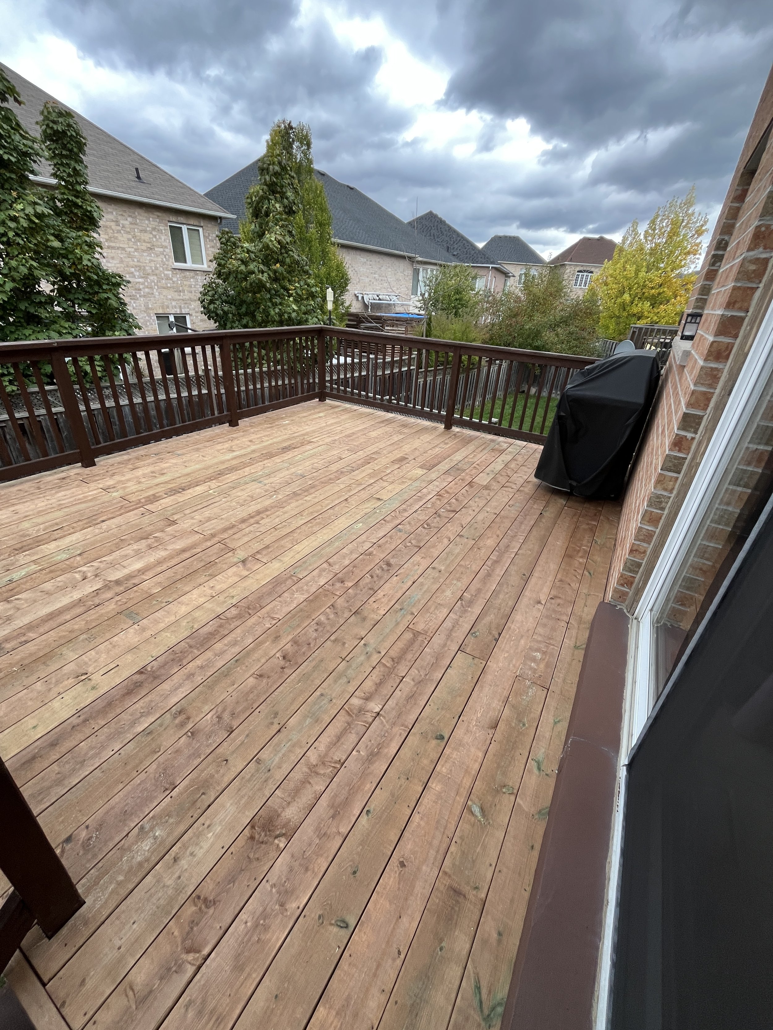 A wooden backyard deck with a railing, partially covered with a black grill cover, and a view of neighboring houses and trees under a cloudy sky.