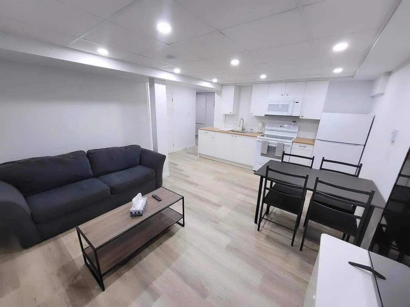 Open-plan living room and kitchen with white cabinets, black dining table and chairs, black sofa, coffee table, and compact appliances. Bright lighting and light wood flooring.