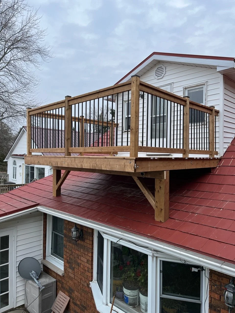 A newly built wooden deck with black metal railing is attached to the second floor of a house with white siding. The house has a red roof, a sliding glass door, and windows. There are leafless trees in the background.