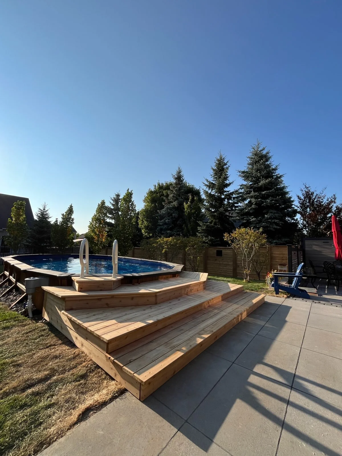 Backyard with a swimming pool on a wooden deck, surrounded by trees and a fence, under a clear blue sky.