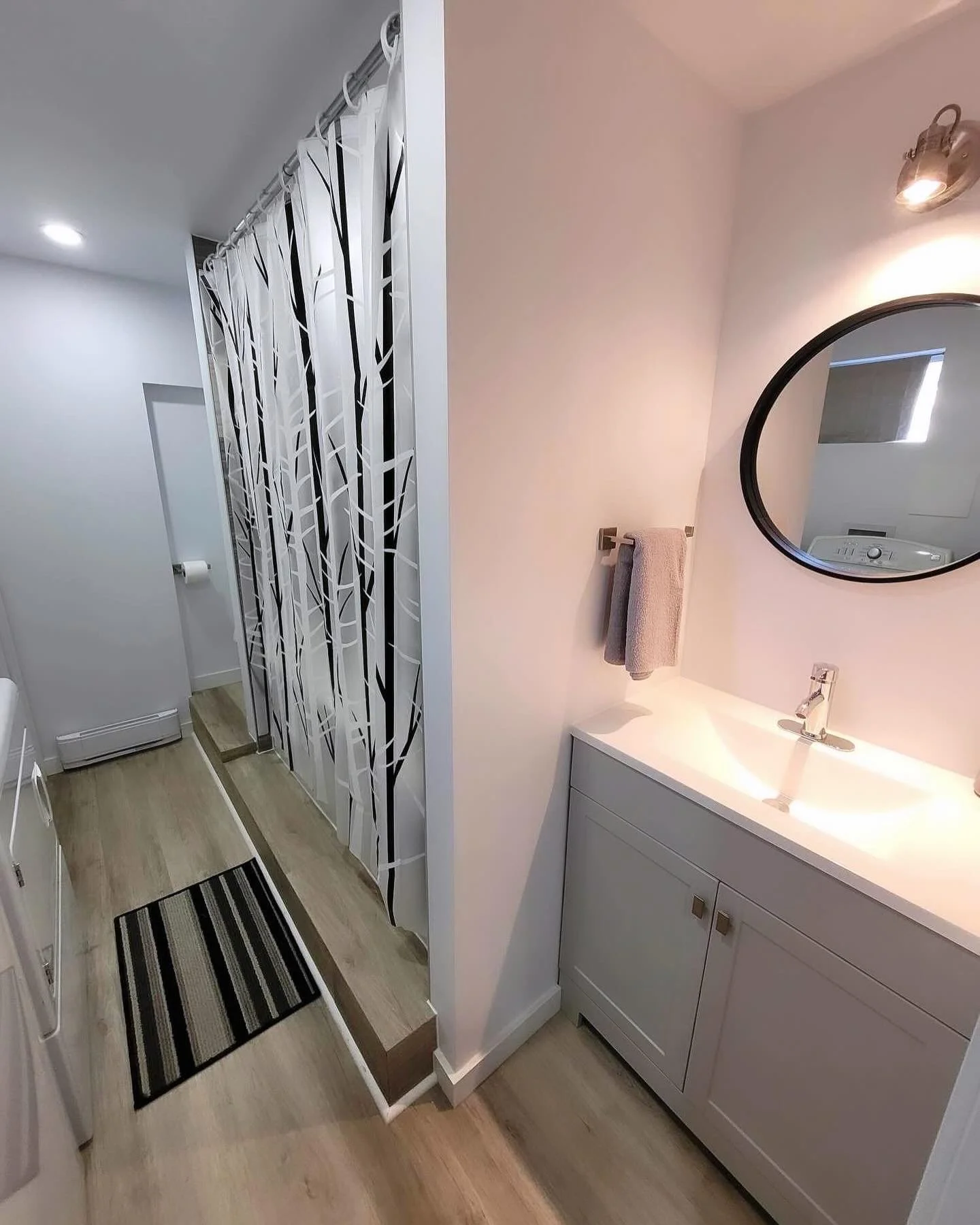 A modern bathroom with a white vanity, a round mirror, a wall-mounted light fixture, a gray towel on a towel bar, and a hallway with a decorative tree-branch patterned privacy screen, a striped rug, and wood flooring.