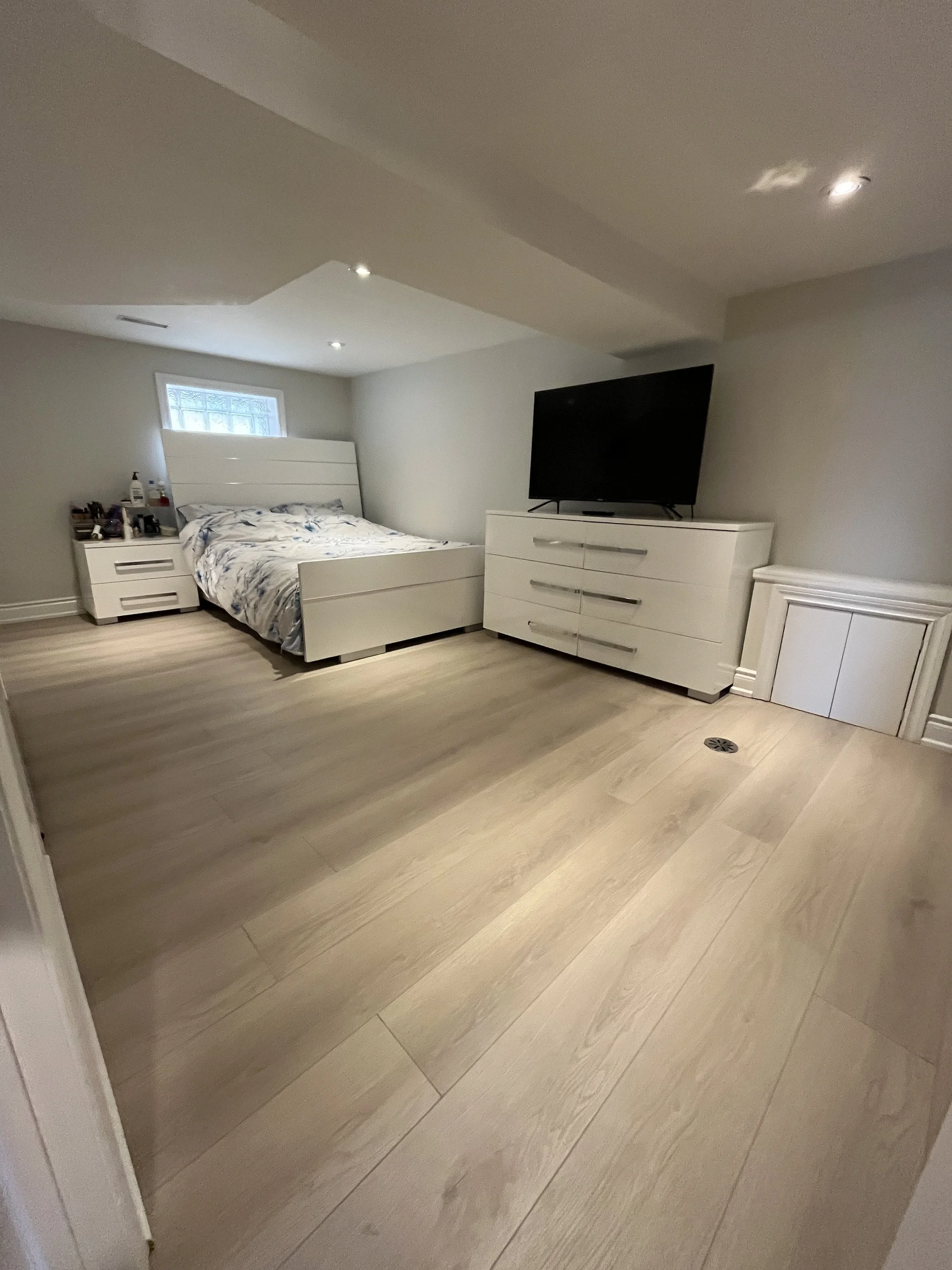 A bedroom with light wood flooring, white furniture including a bed, a dresser with a TV, and a small window above the bed