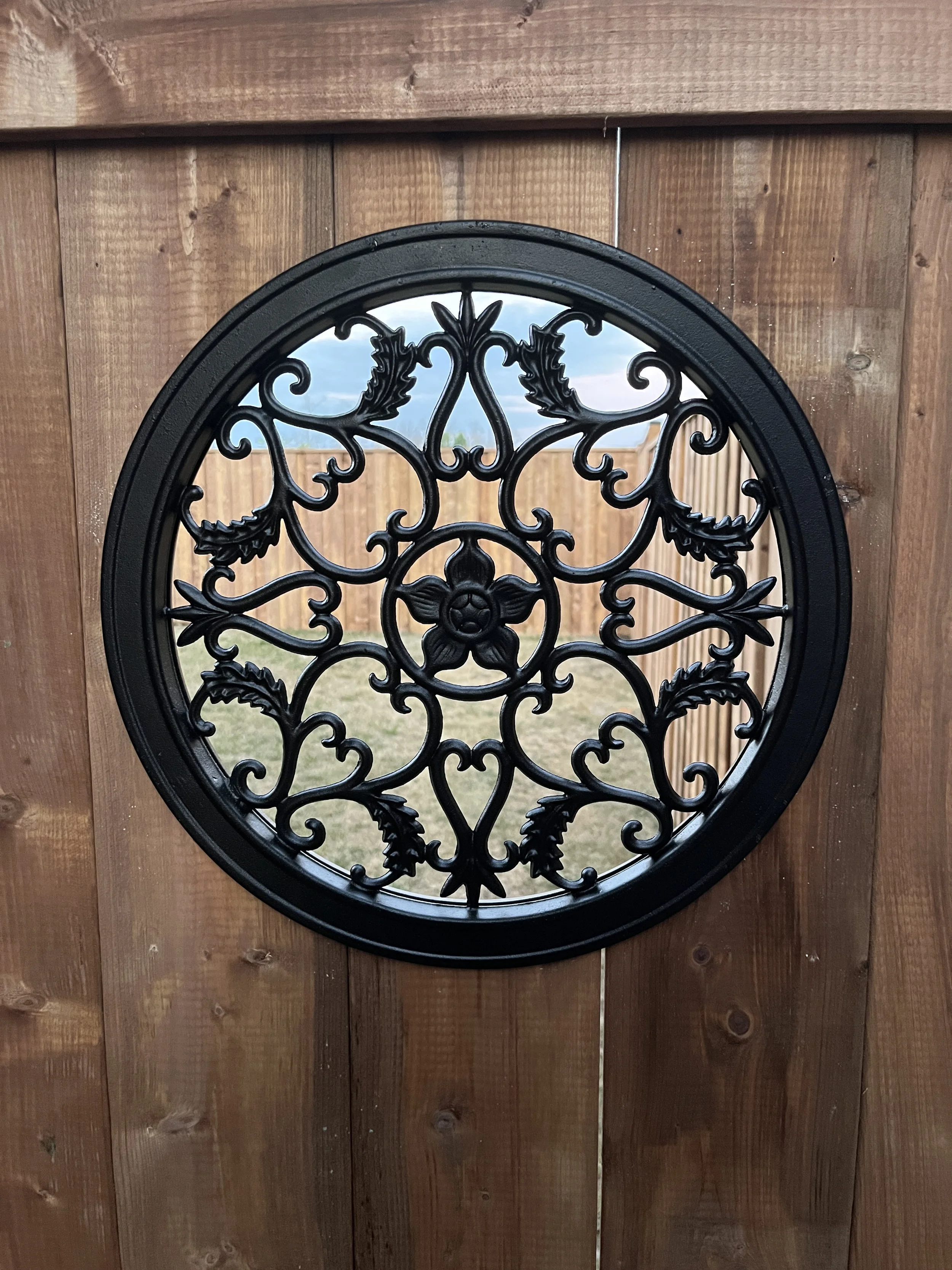 Close-up of a decorative black metal ornament with intricate floral and scrollwork designs mounted on a wooden fence.
