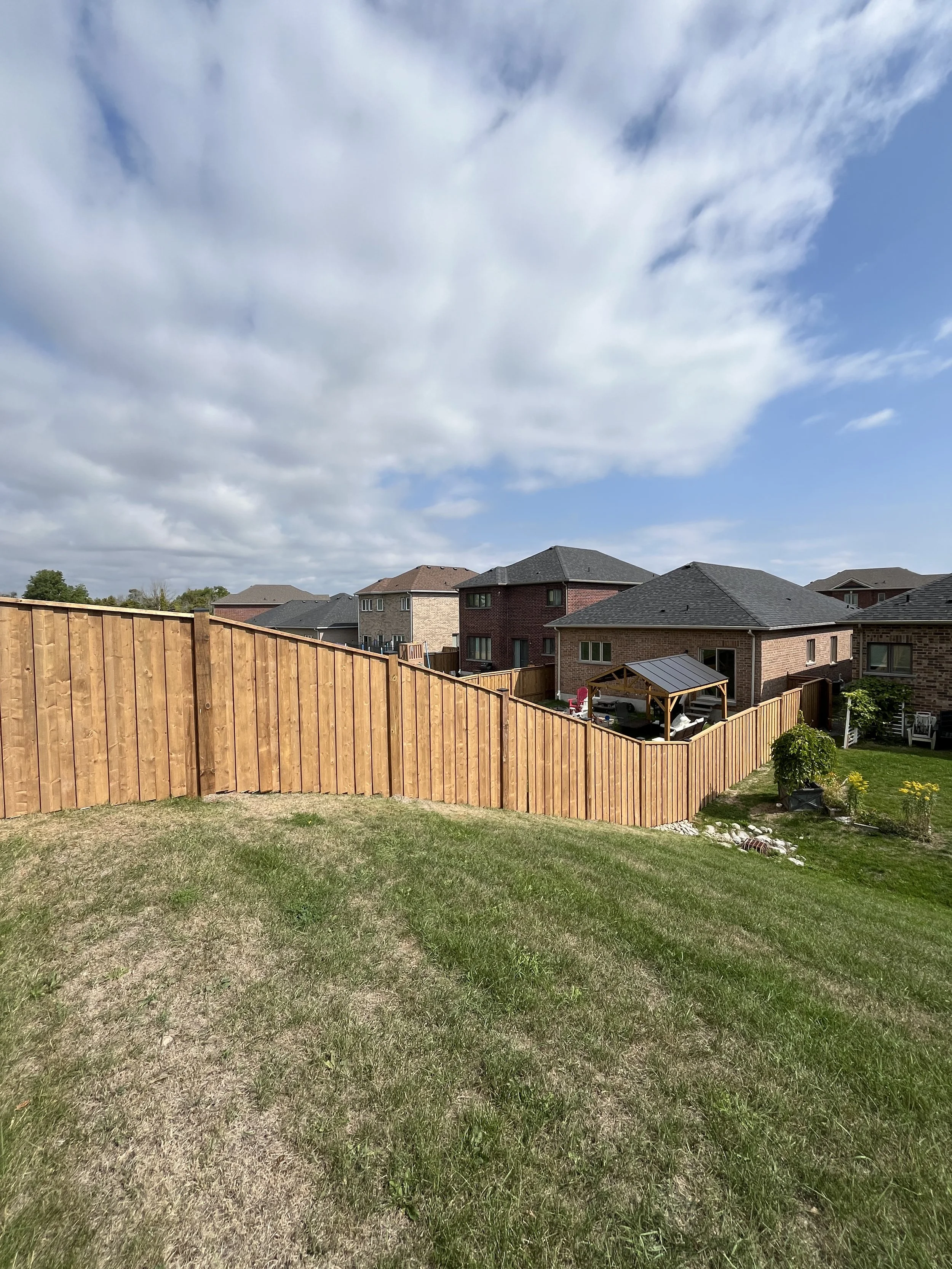 A backyard with a grassy lawn, wooden fence, and neighboring houses with brick and siding exteriors, under a partly cloudy sky.