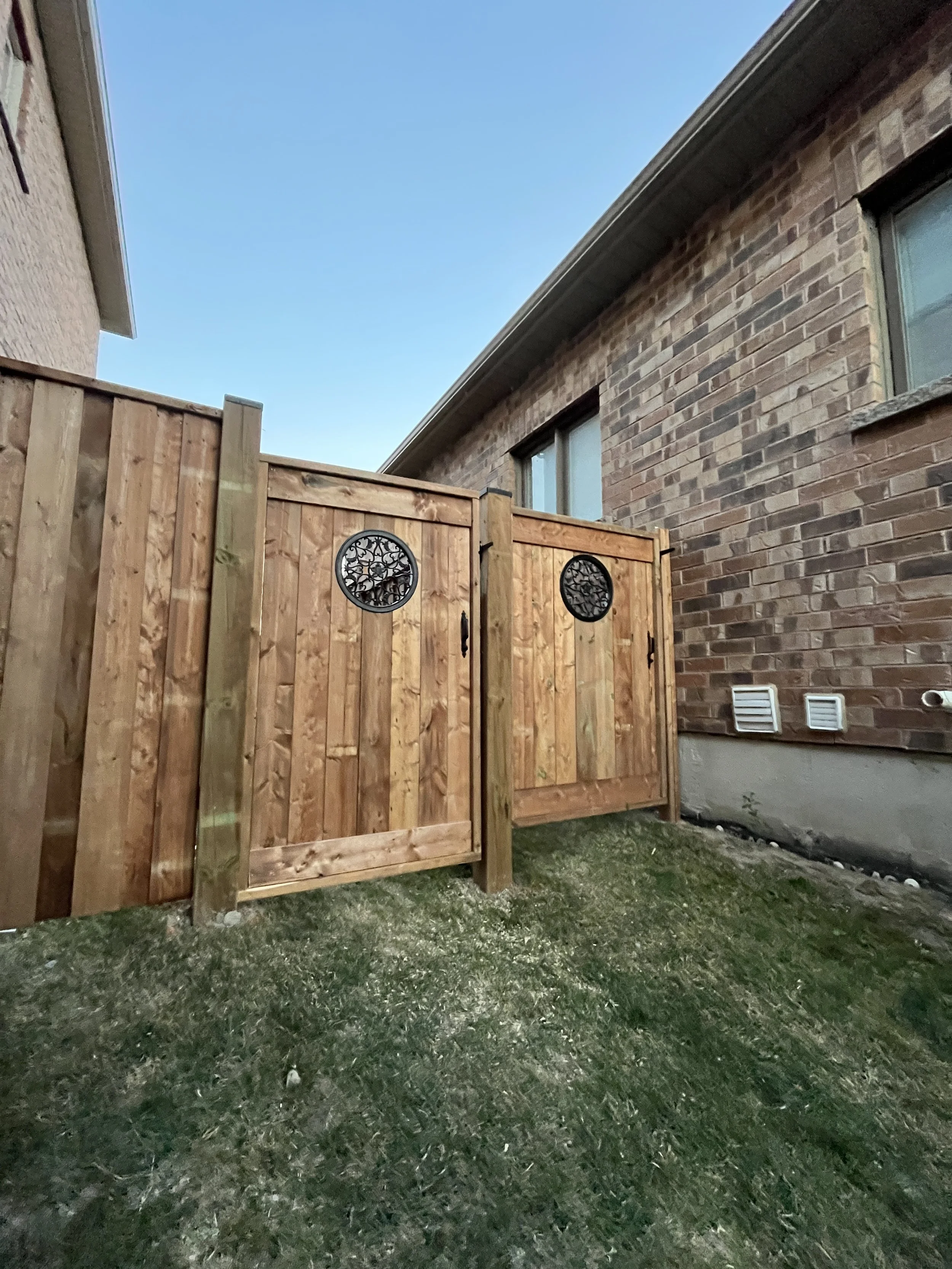 Backyard with a wooden gate in front of a brick house wall, green grass, and a clear blue sky.