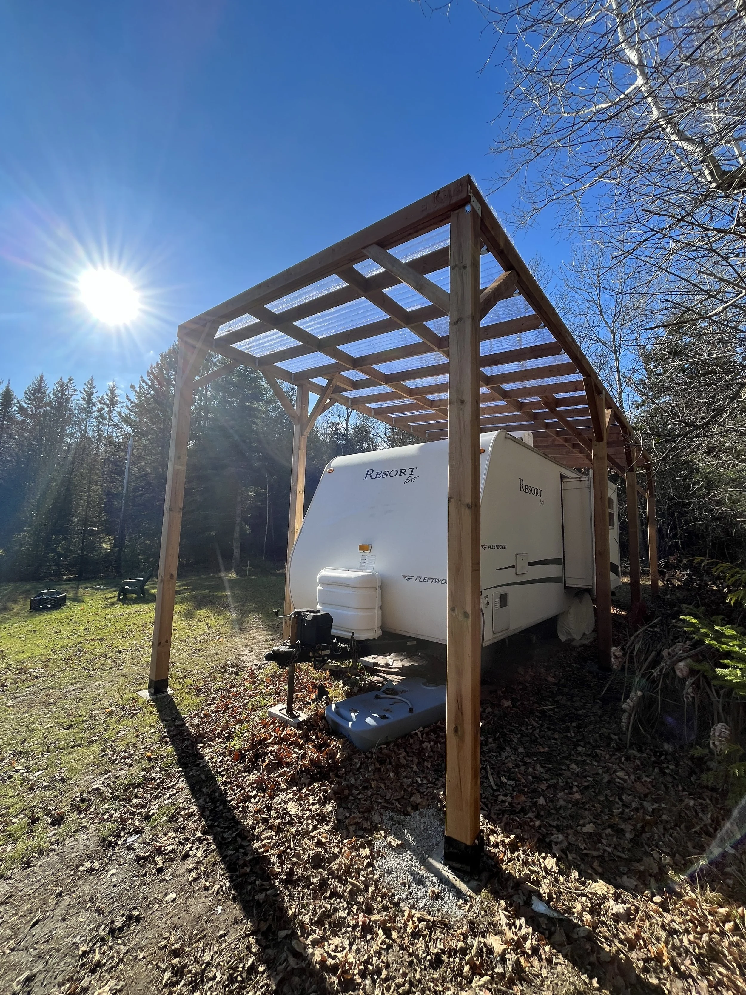 A travel trailer is parked on a patch of grass surrounded by trees with some leaves on the ground. A wooden frame structure is being built over the trailer, likely for a roof or cover. The sky is clear and the sun is shining brightly.