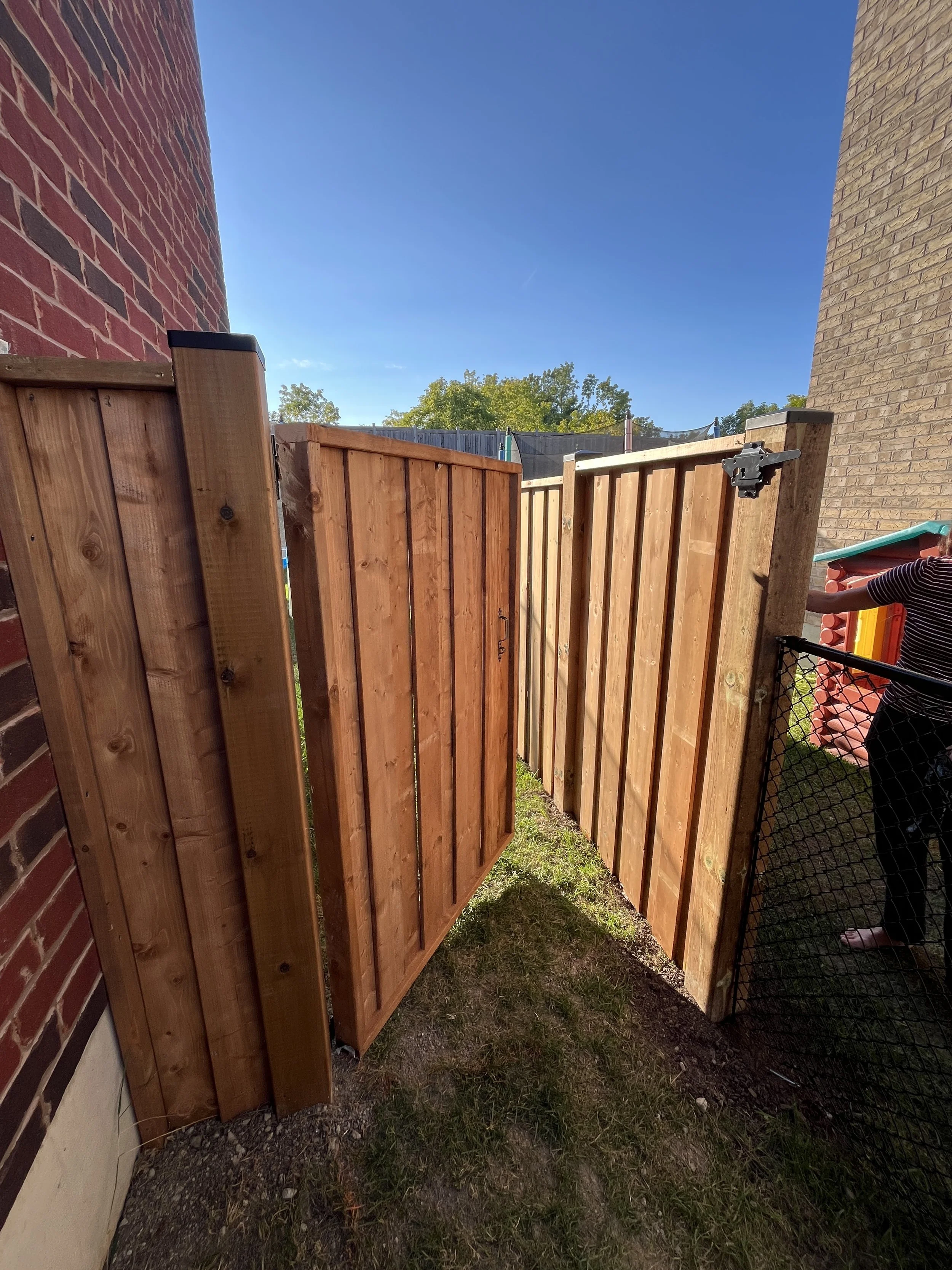 View of a newly installed wooden fence between two brick walls, with a grassy area at the bottom and trees and a blue sky in the background.