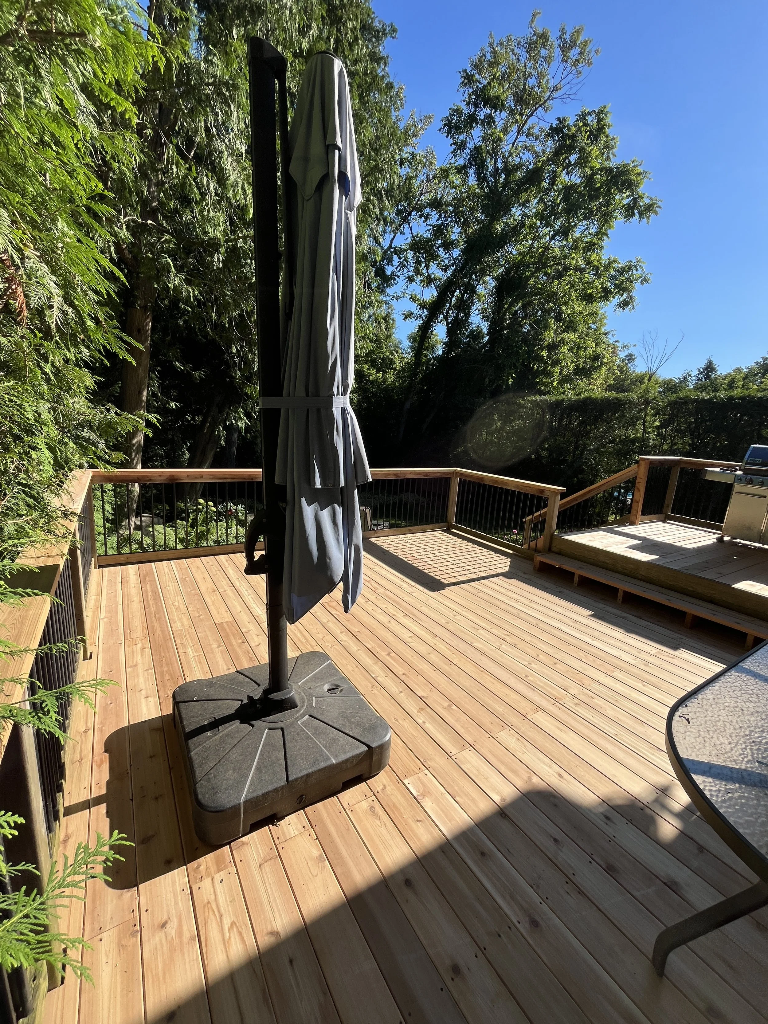 Wooden deck with a closed umbrella, trees and a blue sky in the background, and part of a metal table in the foreground.