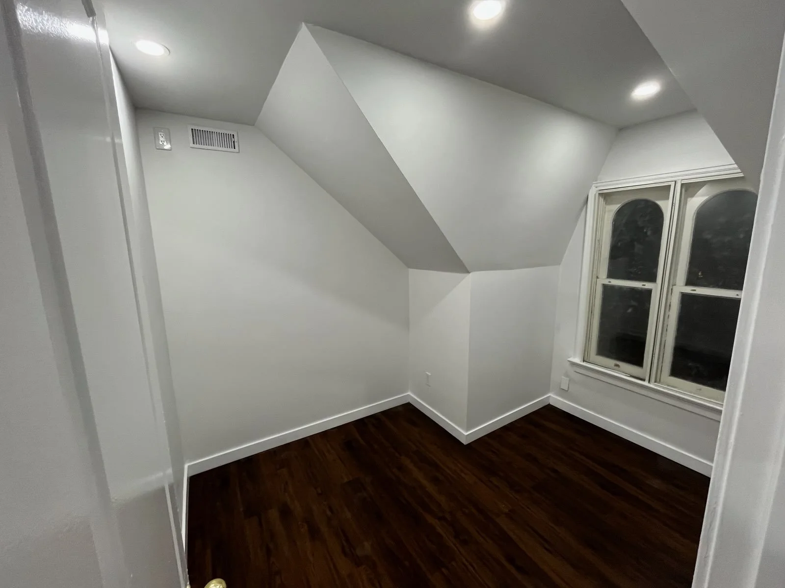 An empty room with white walls, a window with two panes, hardwood flooring, and a ceiling with recessed lighting.