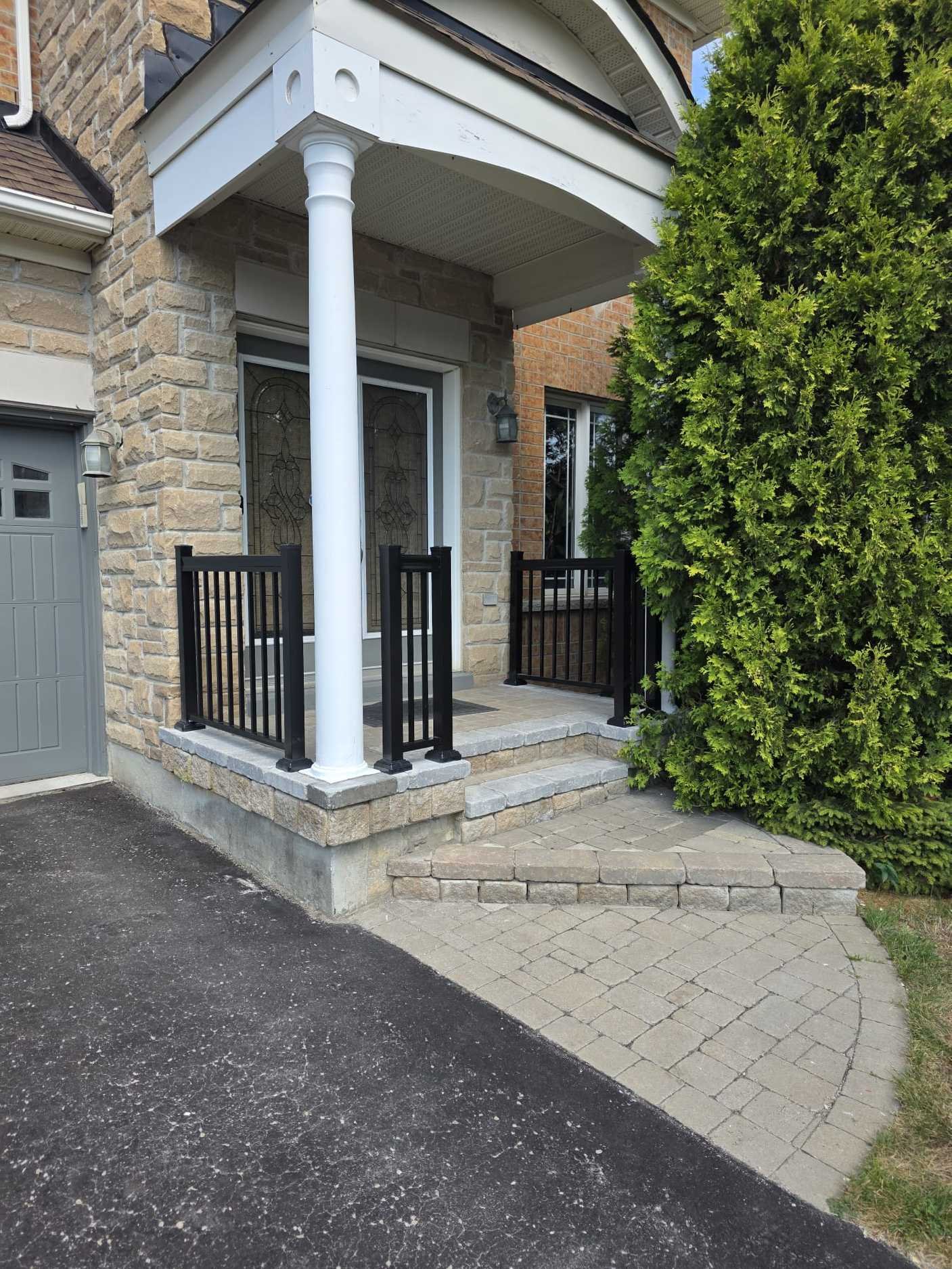 Front porch of a house with a stone step and small steps leading up to the front door, which has glass panels. The porch has black metal railing and a white column supporting the roof. There is a tall green bush on the right side of the porch.