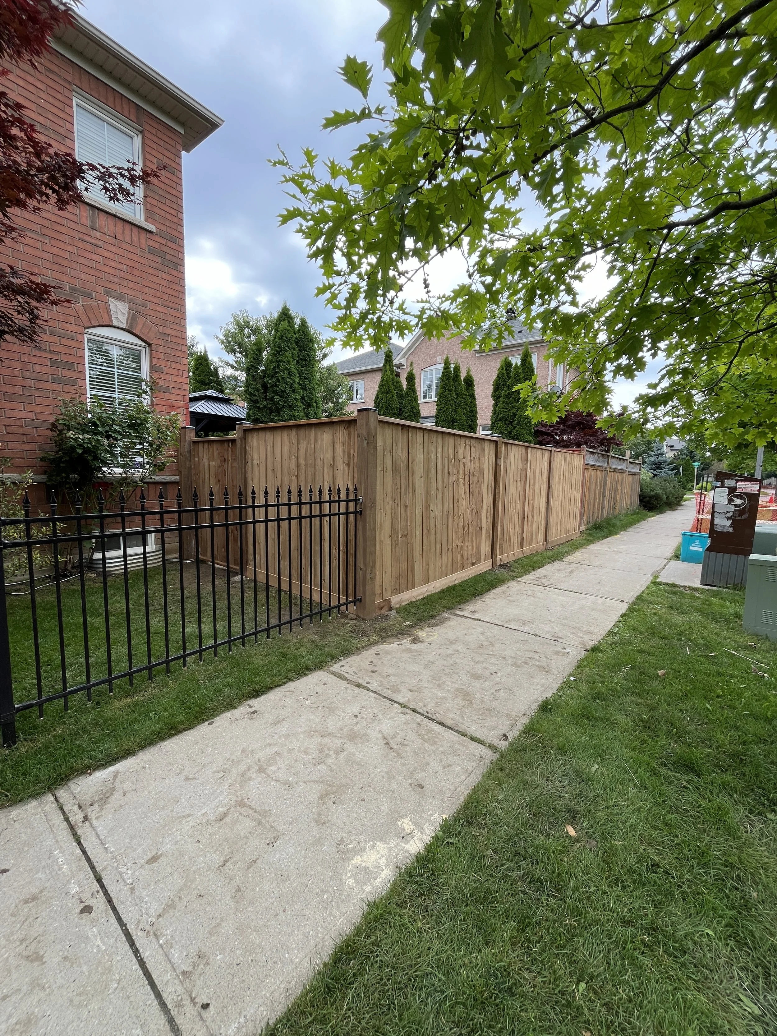 Sidewalk along a grassy yard with a brick house behind a black metal fence and a wooden fence, trees with green leaves, and a cloudy sky.