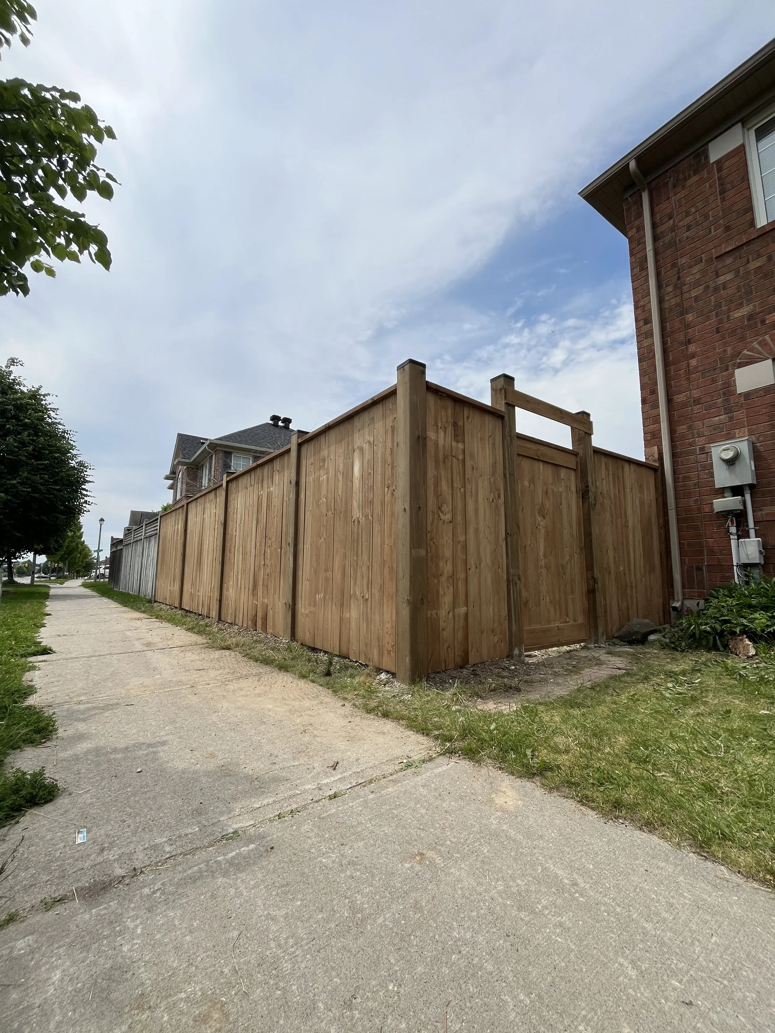 A wooden fence running along a sidewalk next to a brick house, with a cloudy sky overhead and some trees in the background.