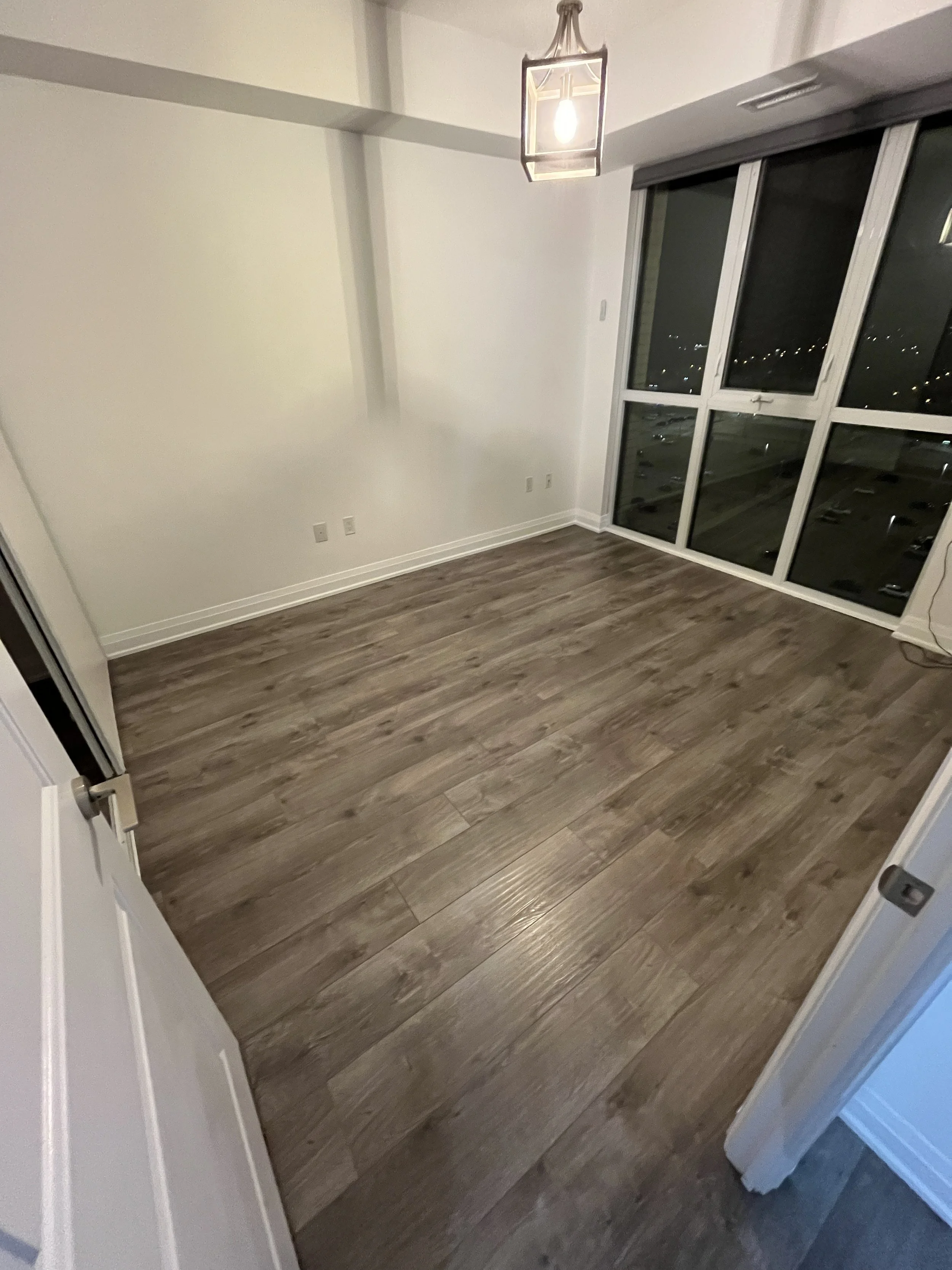 An empty room with wood plank flooring, white walls, a large window with a view of a city at night, and a modern hanging ceiling light fixture.