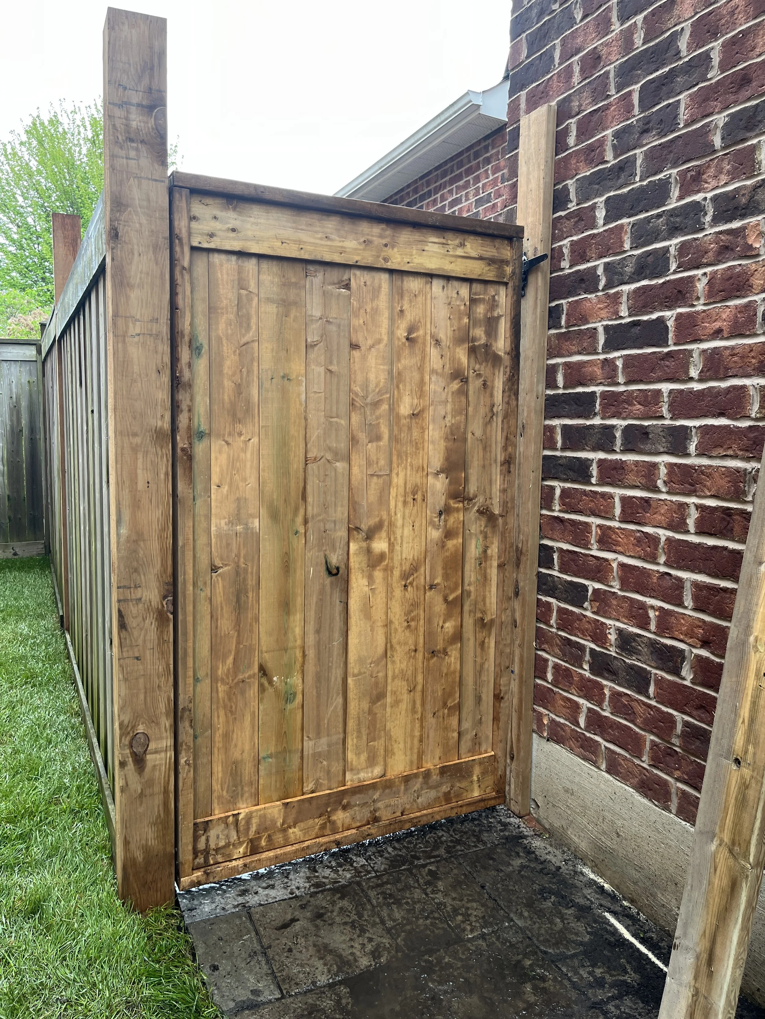A newly installed wooden outdoor shower with a brick wall on one side and grass on the other, showing a black metal handle on the shower door.