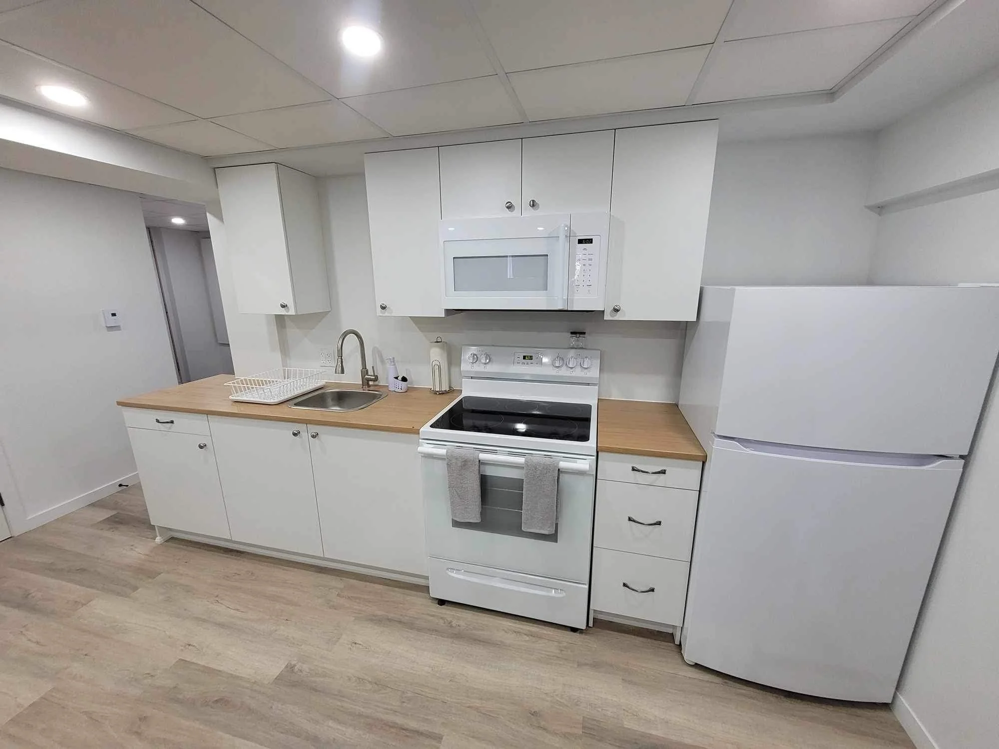 A modern kitchen with white cabinets, a microwave installed above a stove, a stainless steel sink, and a white refrigerator. The kitchen has a light-colored wood floor and a beige countertop.