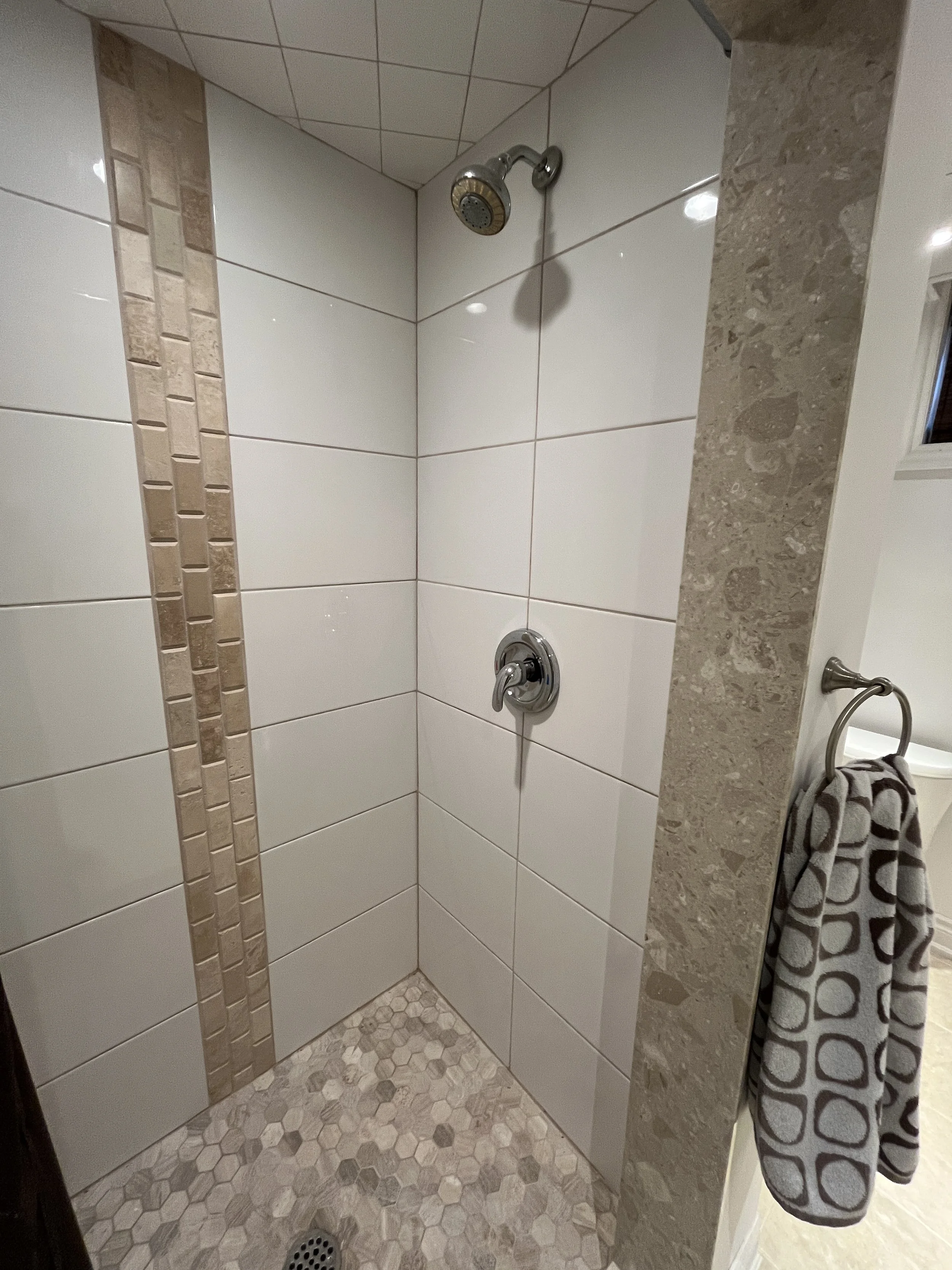 Bathroom shower area with white tiled walls, a beige partial tile border, a showerhead, a faucet, and a hand towel hanging on a hook.