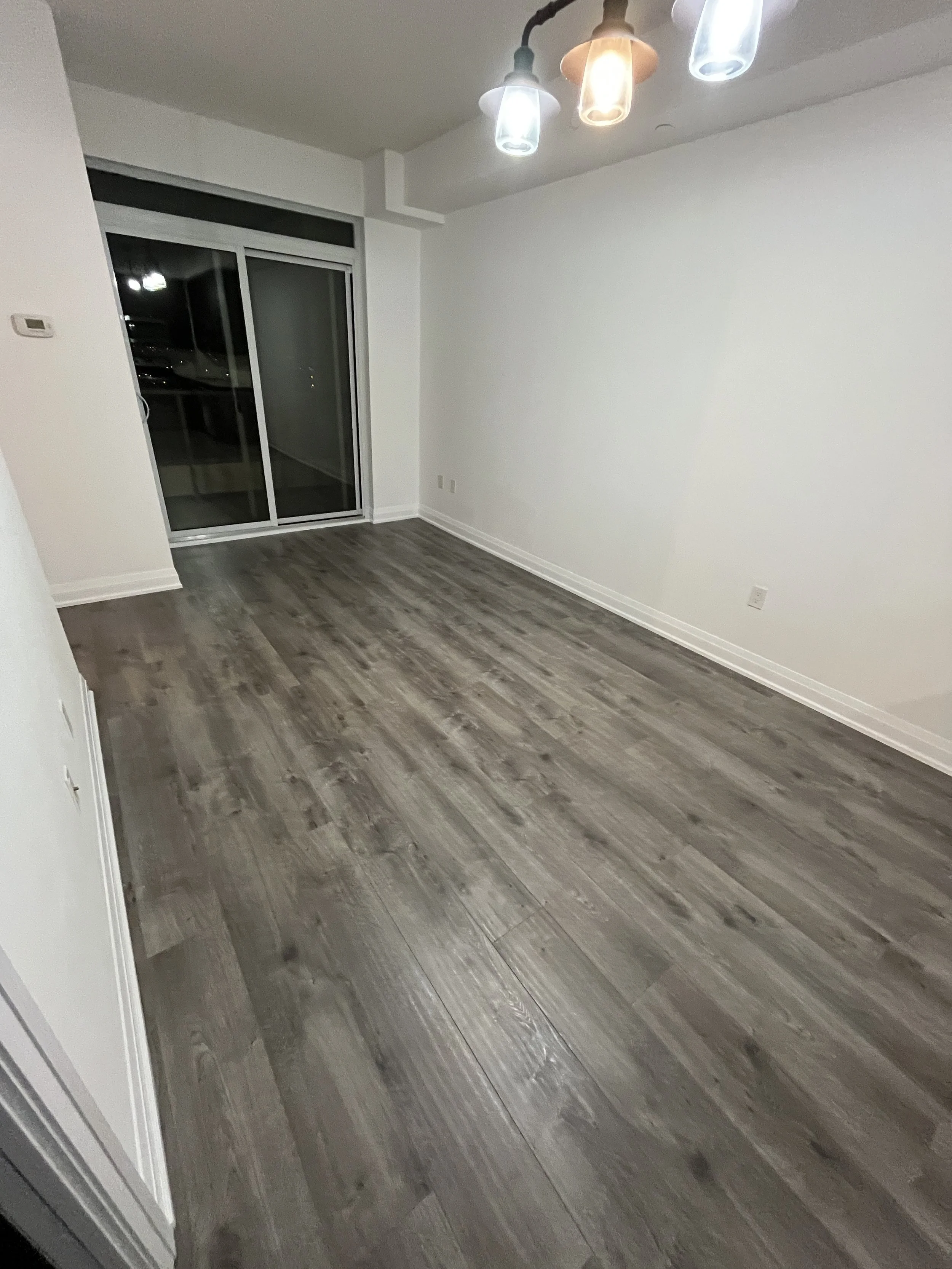 Empty room with wood flooring, white walls, sliding glass door leading to a balcony, and hanging ceiling lights.