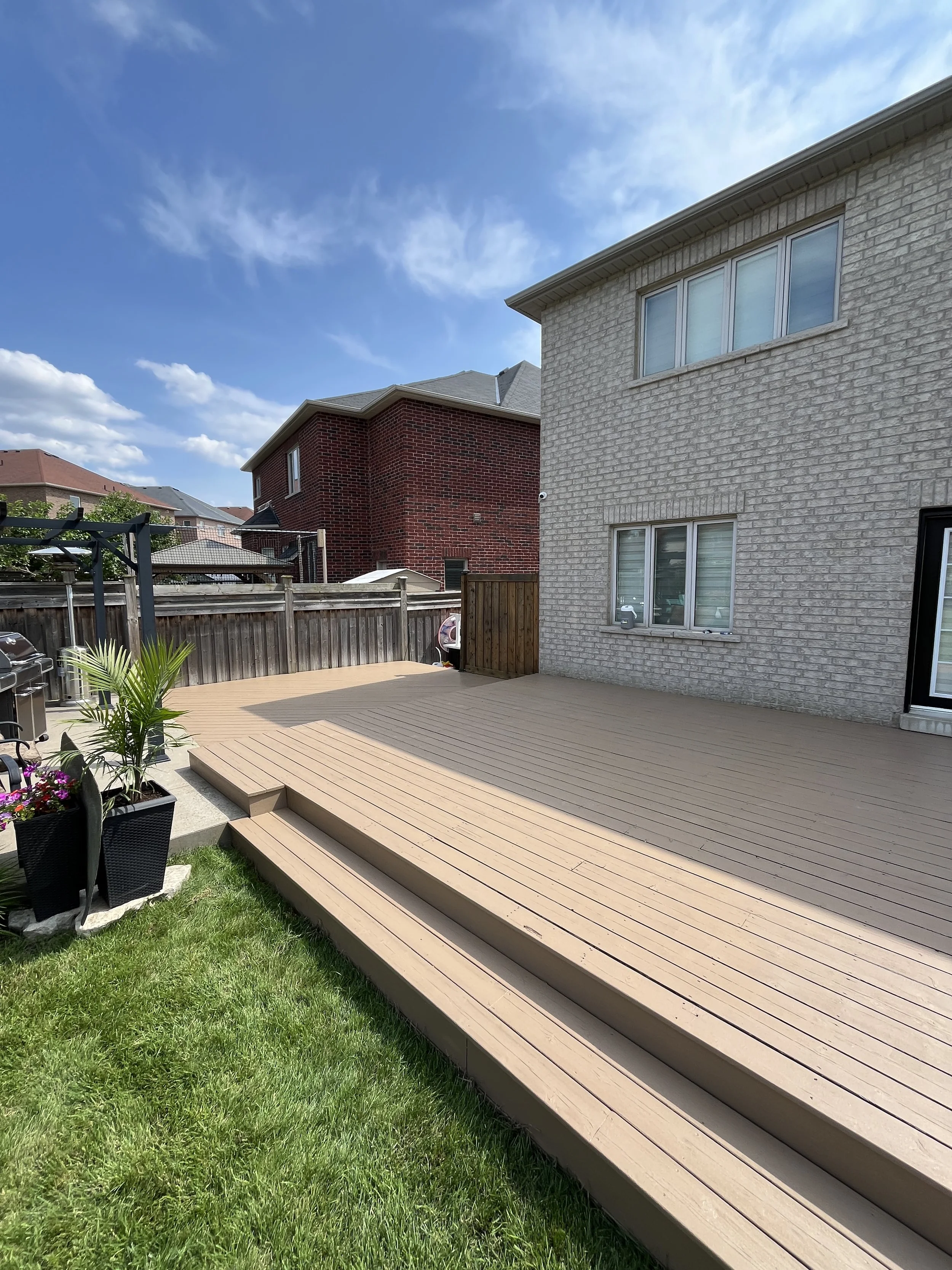 Backyard with a wooden deck, potted plants, and a grassy lawn. Fences and neighboring houses visible under a partly cloudy sky.