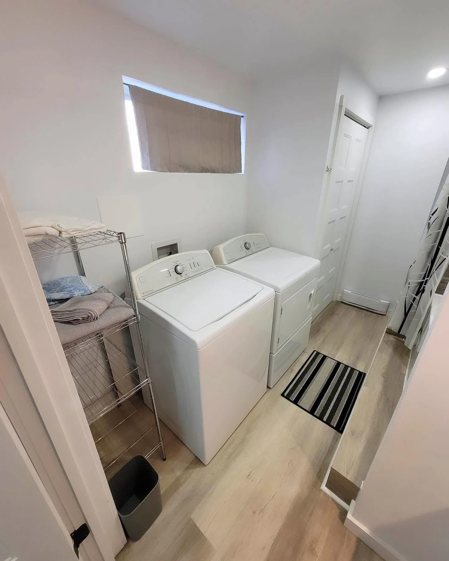 Laundry room with white washer and dryer, metal shelving with folded towels, small window covered with a brown blind, striped rug, and a white door.