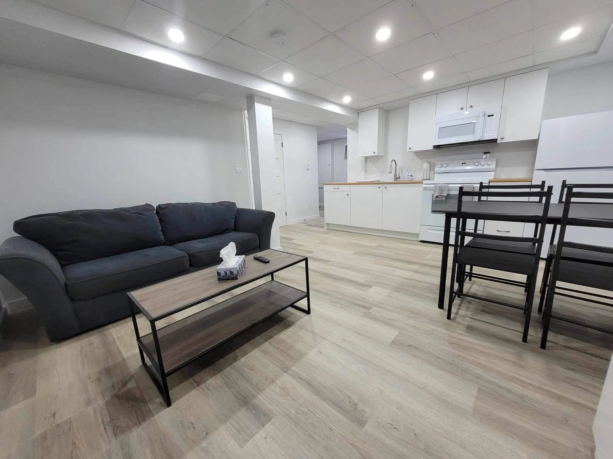 Living room area with black sofa, coffee table, and remote control. Open kitchen with white cabinets, microwave, stove, and refrigerator. Dining table with black chairs. Light hardwood flooring and ceiling with recessed lighting.