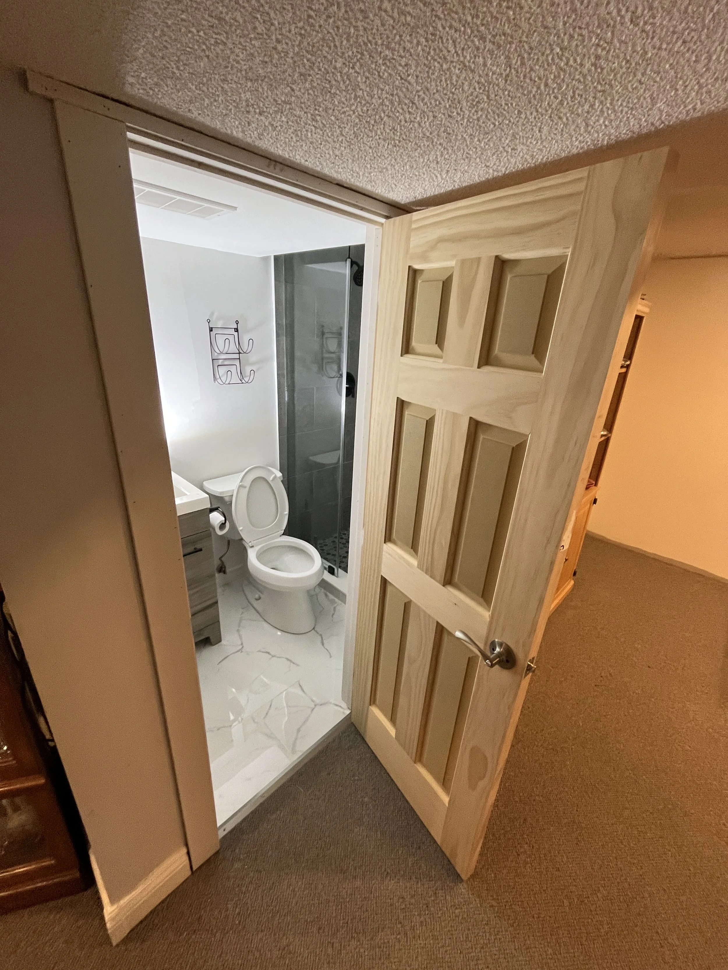 A partially opened wooden door reveals a bathroom with a toilet, a window, and a shower stall with glass door, white walls, and grey tile.