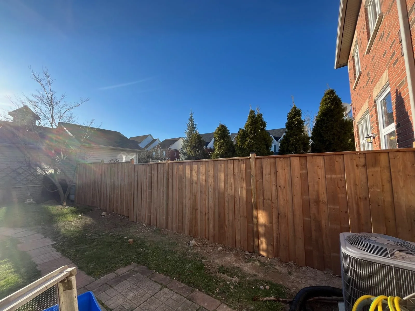 Backyard with new wooden fence, neighboring houses, trees, and clear blue sky.