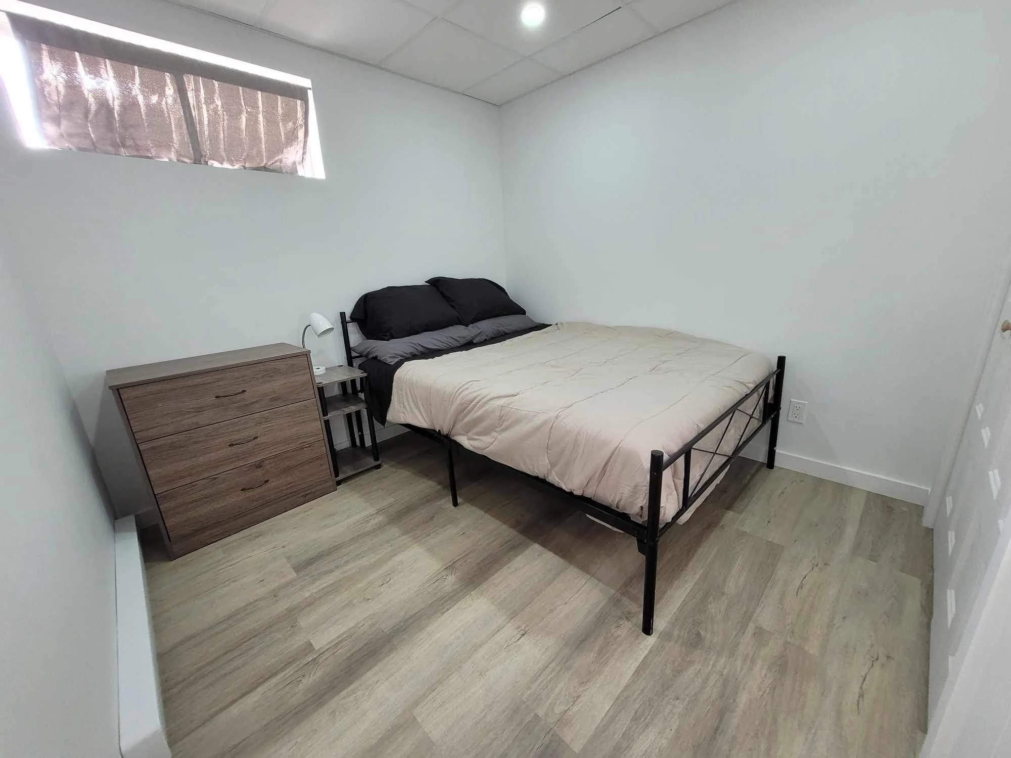 A simple bedroom with a single bed, black and gray pillows, a wooden nightstand with a small white lamp, and a window with a partially pulled curtain. The walls are white, and the flooring is wood.