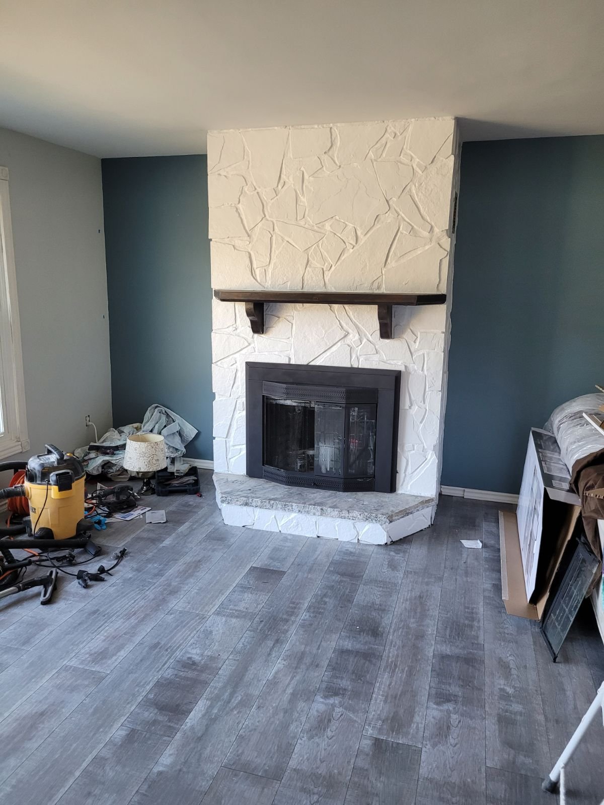 Living room with a white stone fireplace, surrounded by construction tools and materials, with blue and gray walls and wooden flooring.