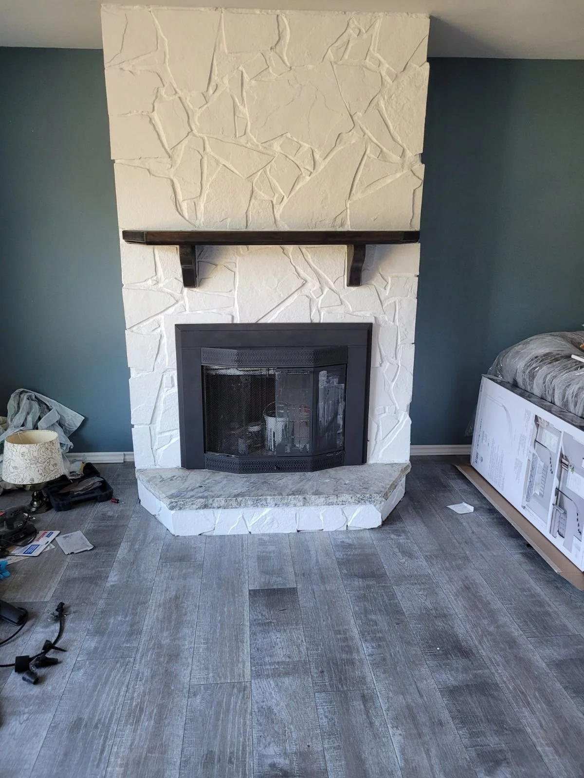 Living room with a white textured stone fireplace, black mantel, dark wood floor, and a bed with plastic covering on the right side. Construction tools and supplies are scattered on the floor.