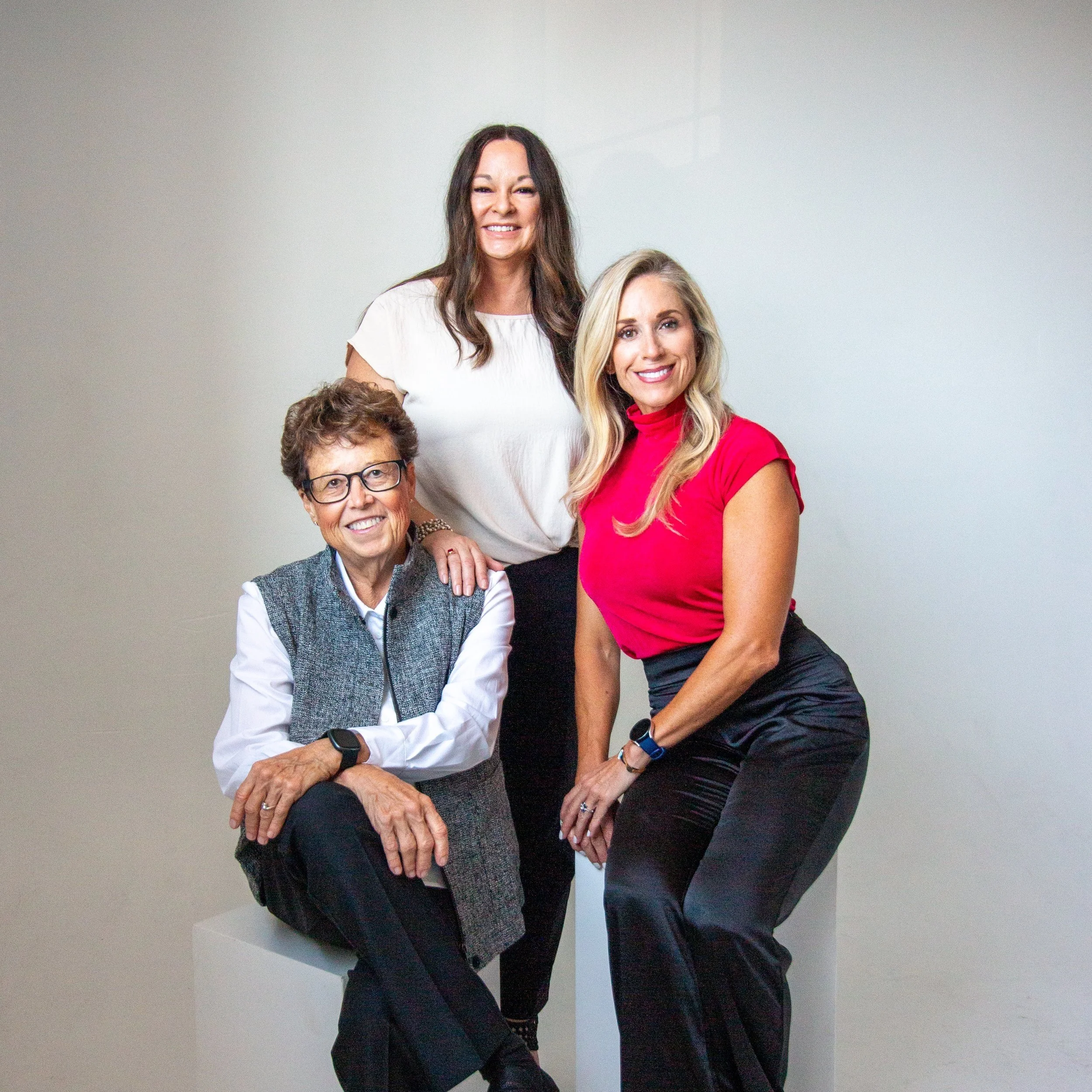 Three women posing for a group photo in a studio setting. The woman on the left is seated wearing glasses, a white shirt, and a gray vest. The woman in the middle is standing, dressed in a white top and black pants. The woman on the right is seated, wearing a red sleeveless top and black pants.