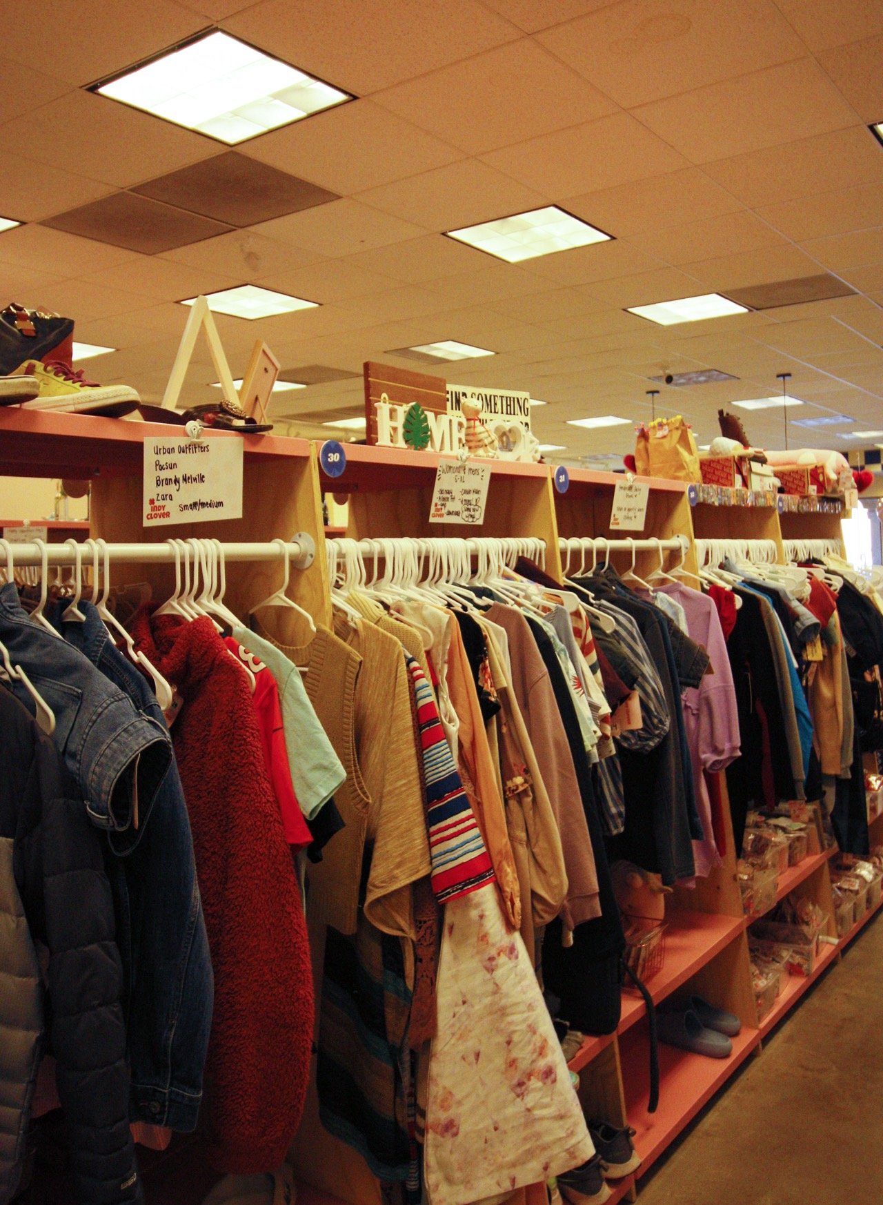 Interior of a thrift store with clothing racks displaying various garments, shoes on top shelves, and items in bins below.