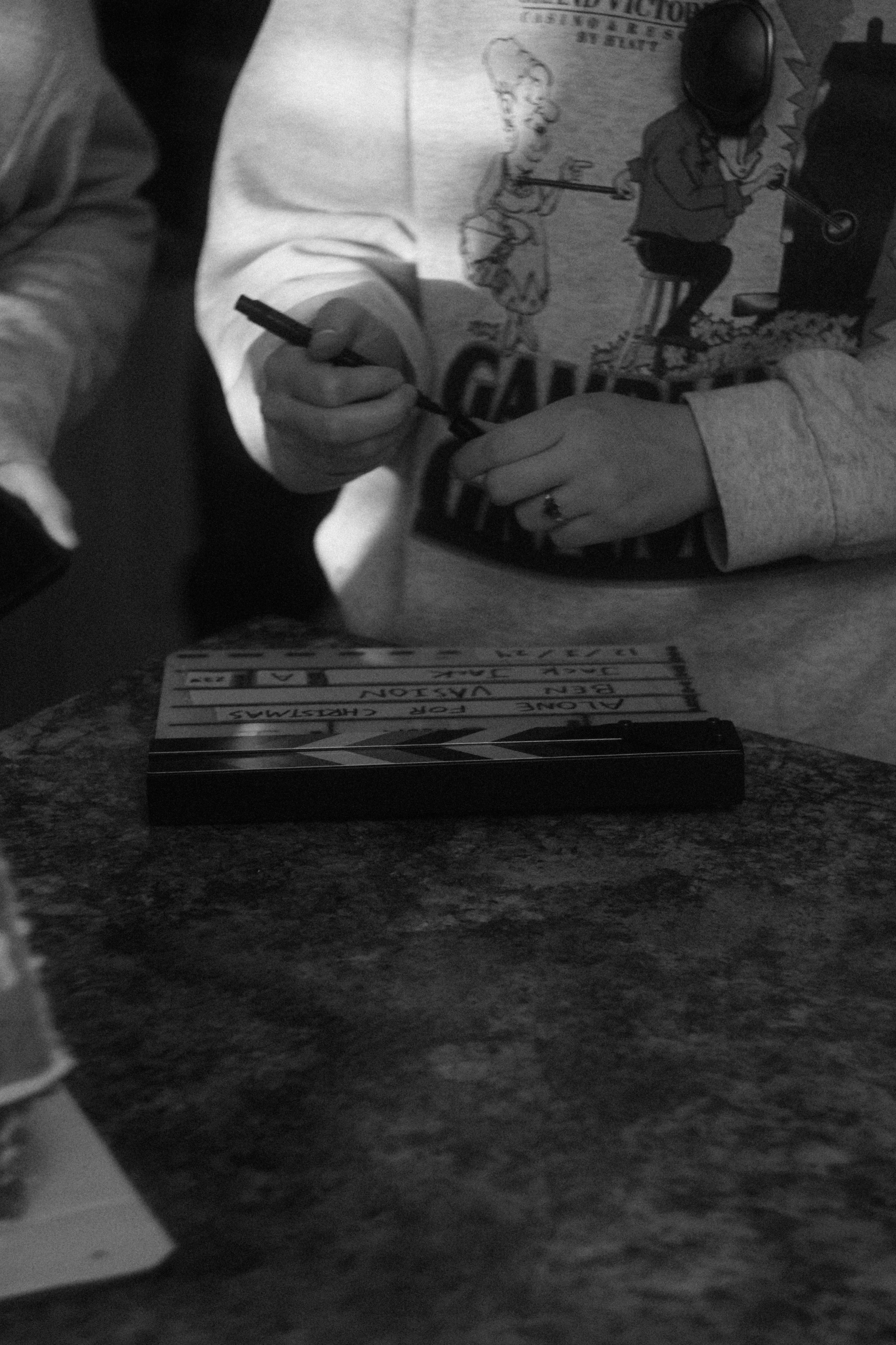 Black and white photo of a person in a cartoon-print sweatshirt writing on a film clapperboard with a marker.