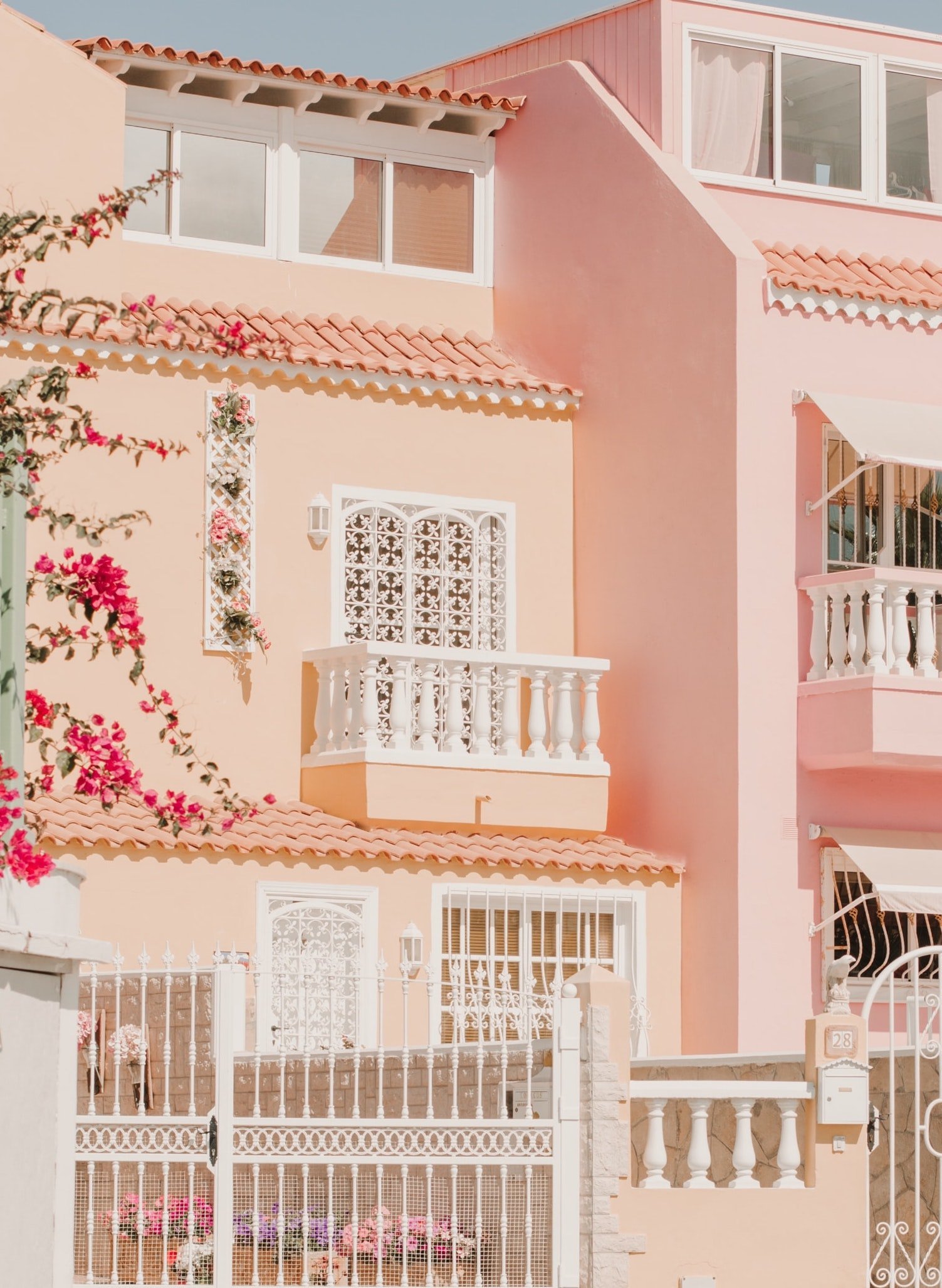 Pink residential building with white decorative railings, a gated entrance, and pink flowering plants outside.