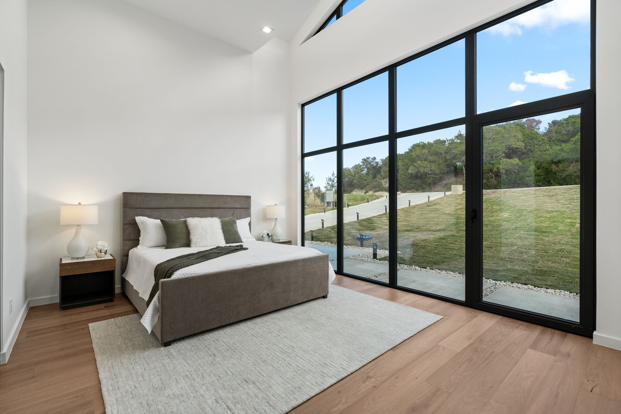 Modern bedroom with large window, gray bed, white bedding, wooden floors, and minimalist decor.