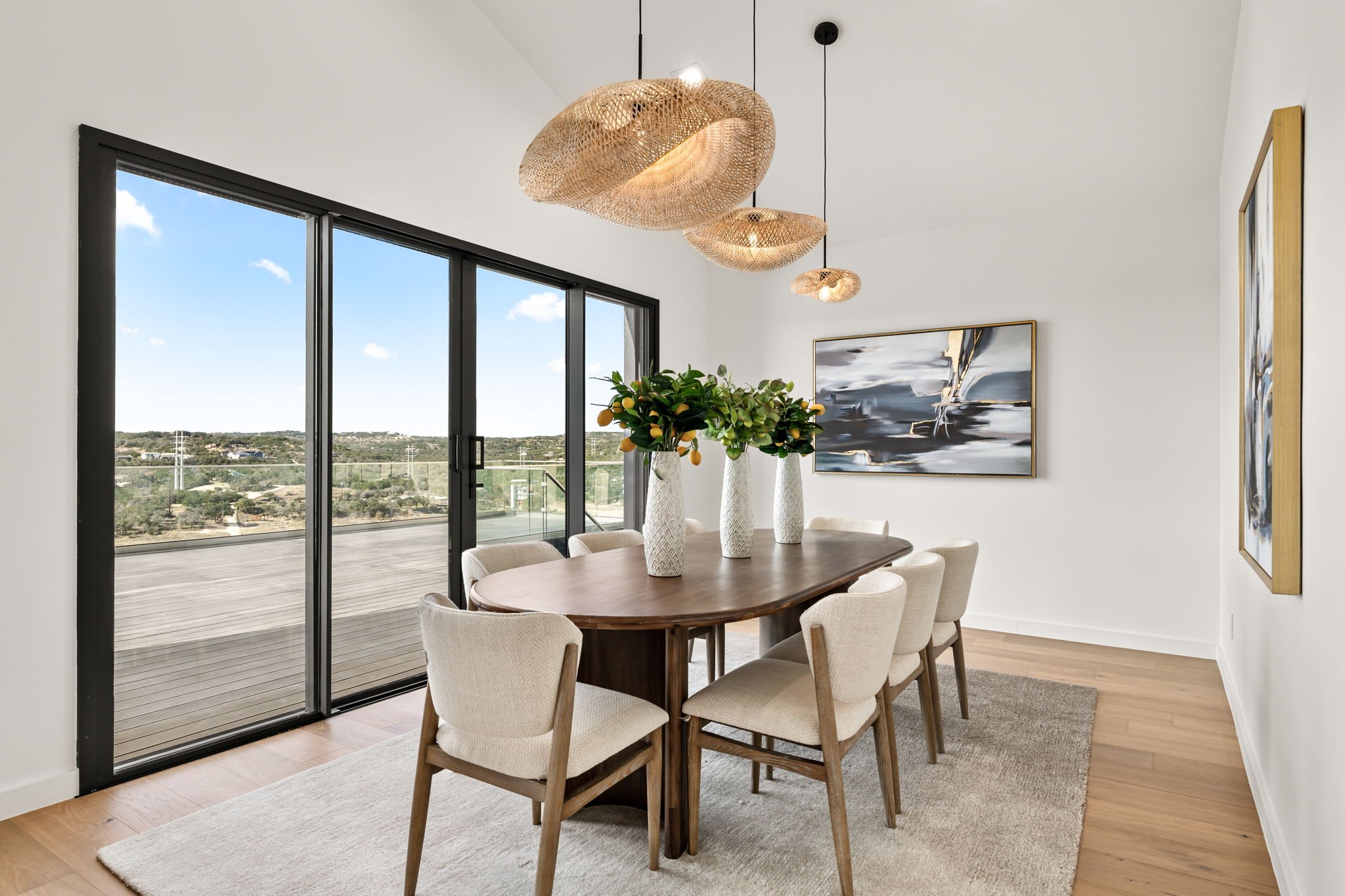 Modern dining room with a wooden table, beige upholstered chairs, large abstract artwork, pendant lights, and a view of a balcony through glass doors.