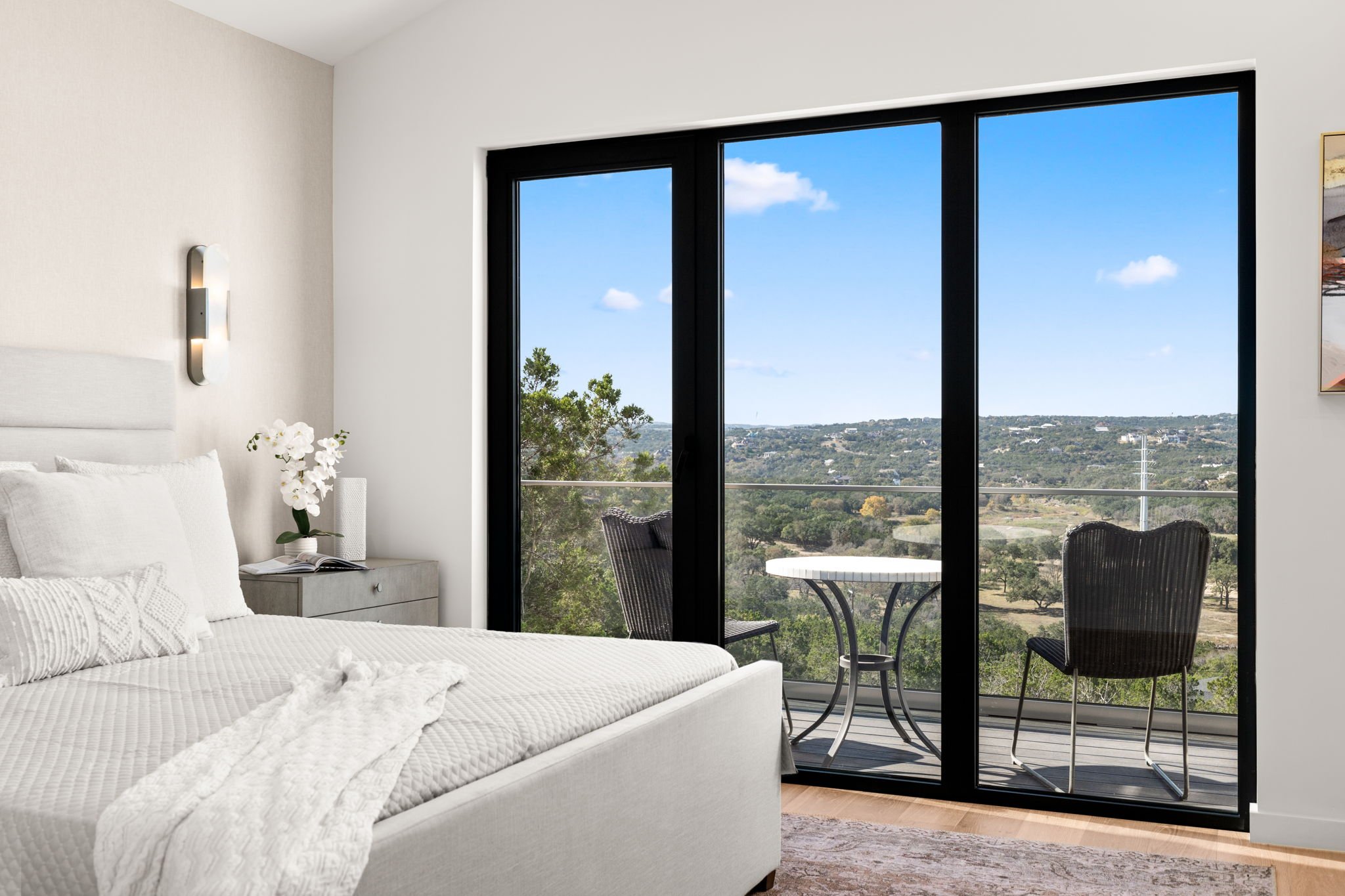 Modern bedroom with a large bed, bedside table with a vase of white flowers, and a sliding glass door leading to a balcony with a small table and chairs overlooking a scenic view.
