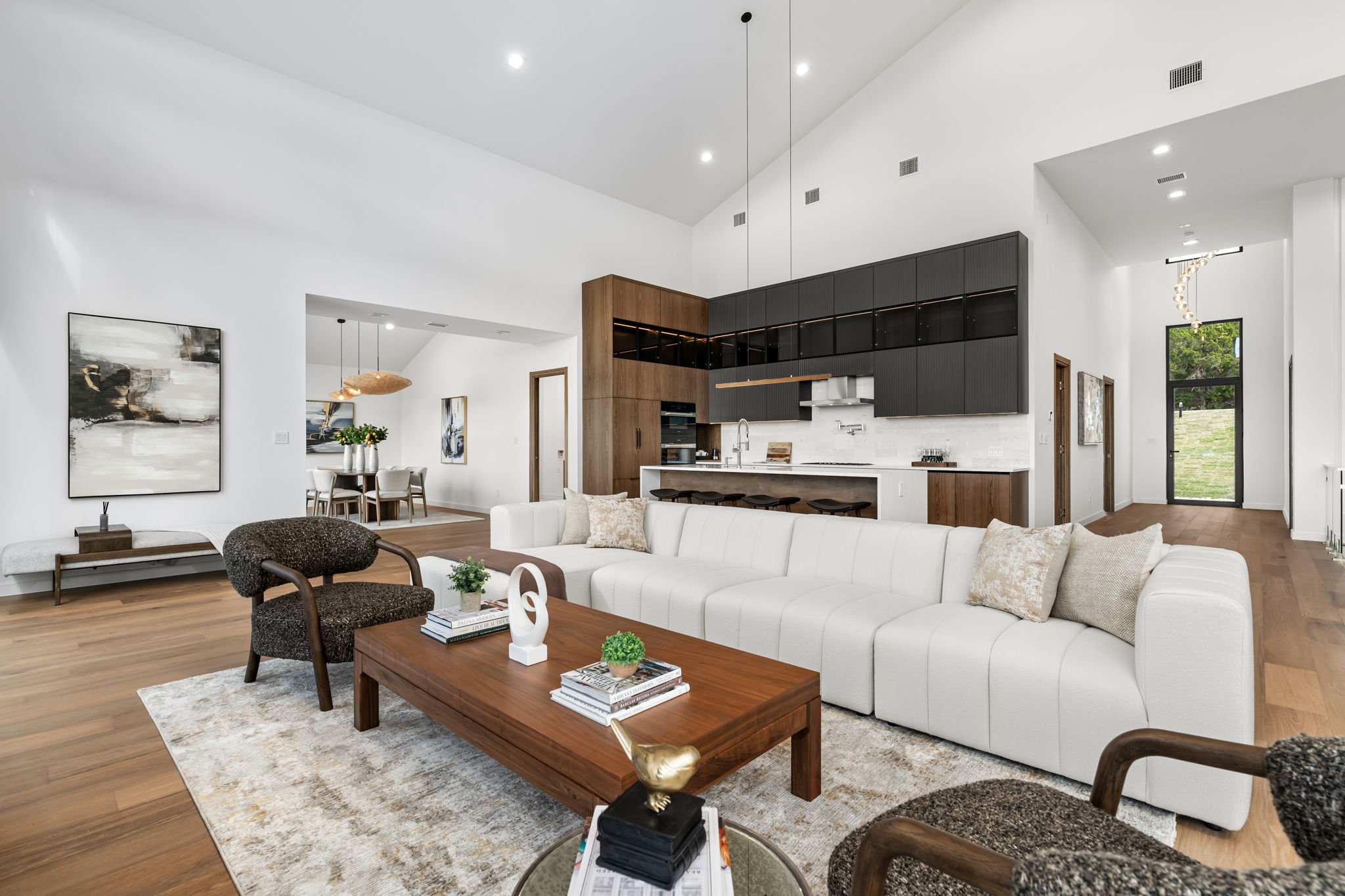 Modern open-concept living room with a white sectional sofa, wooden coffee table, contemporary armchairs, and a kitchen in the background. The space features high ceilings, recessed lighting, and minimalist decor with neutral tones.