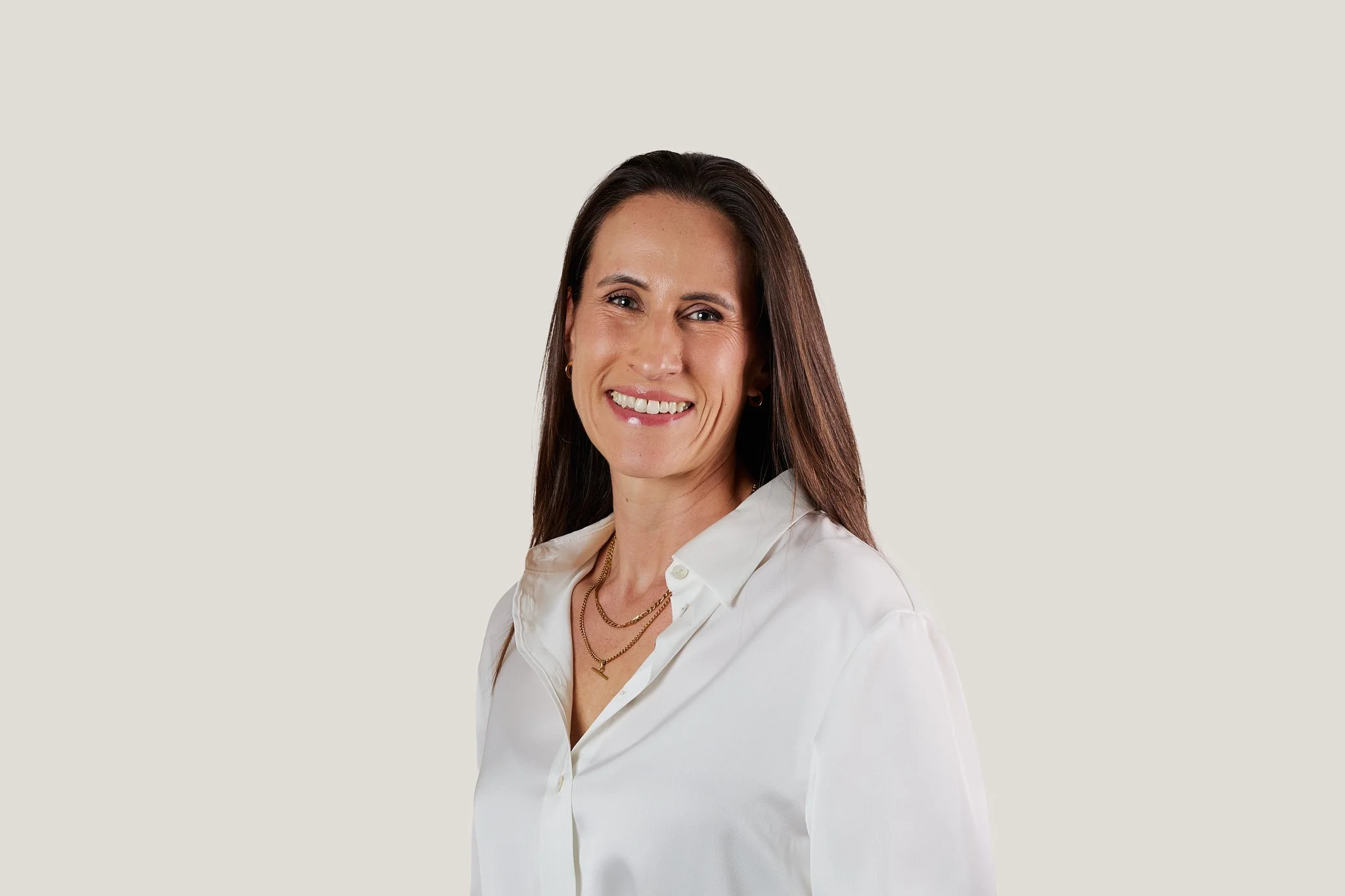 A woman with long brown hair wearing a white button-up shirt, gold jewelry, and smiling.