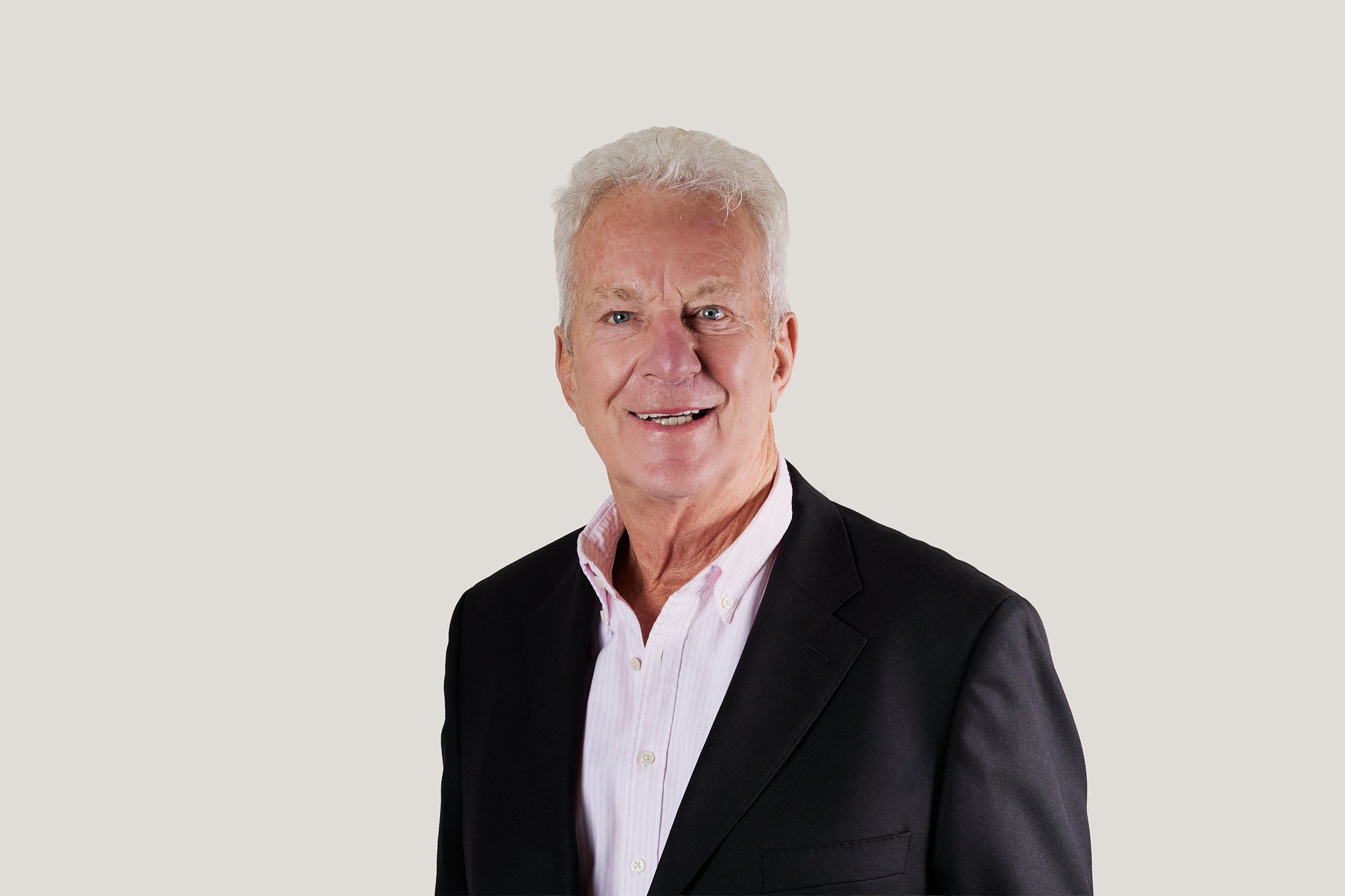 An older man with gray hair, wearing a black blazer over a pink striped shirt, smiling against a plain off-white background.