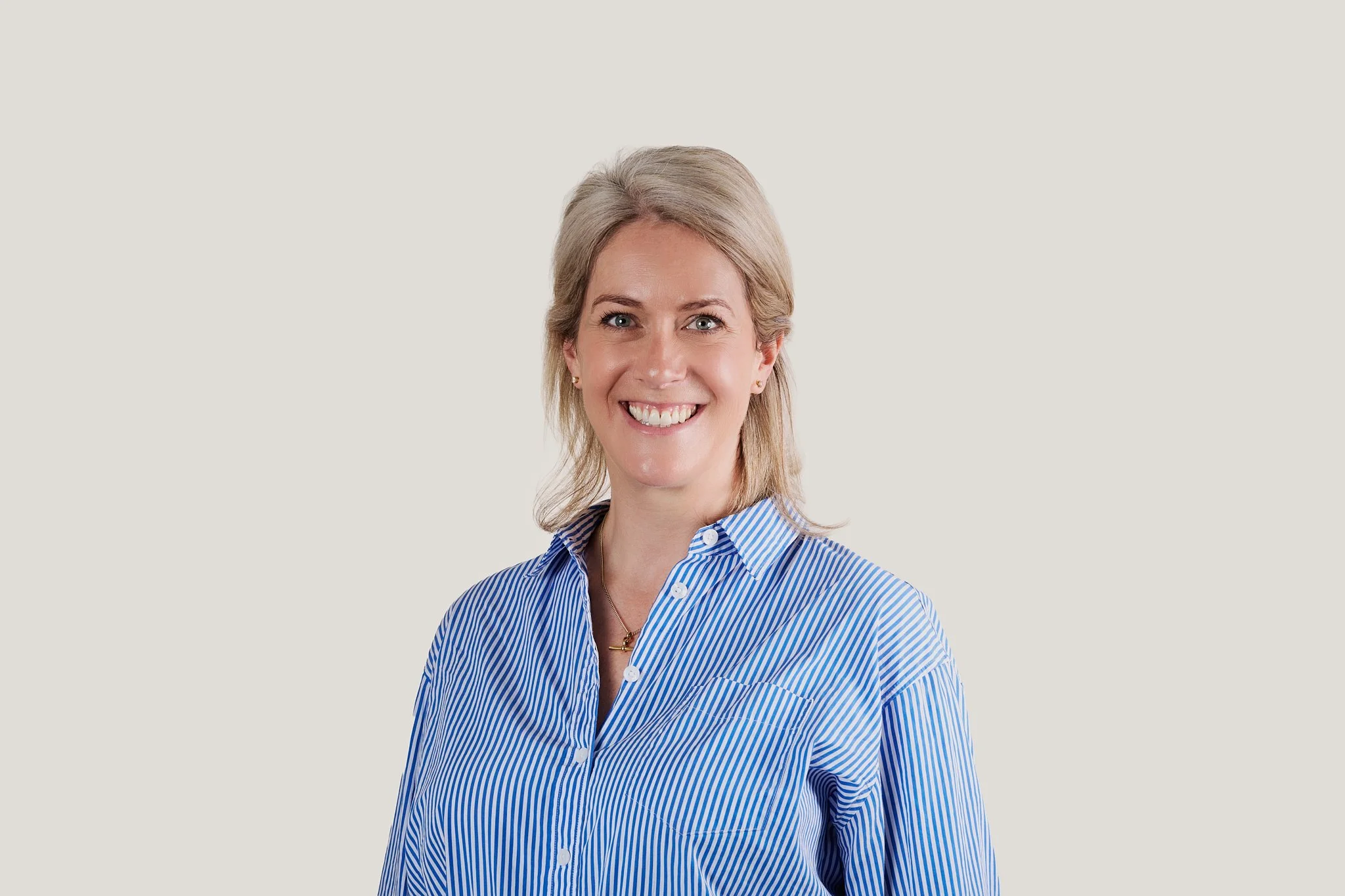 A smiling woman in a blue and white striped button-up shirt with blonde hair styled in a layered cut against a plain, light-colored background.