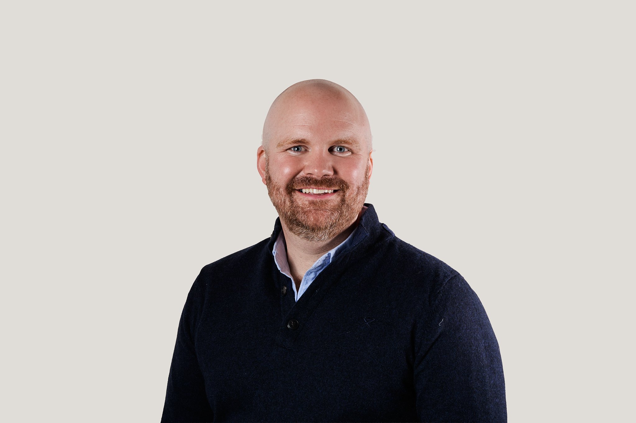 A smiling man with a bald head and beard, wearing a dark sweater and light collared shirt, against a plain light background.