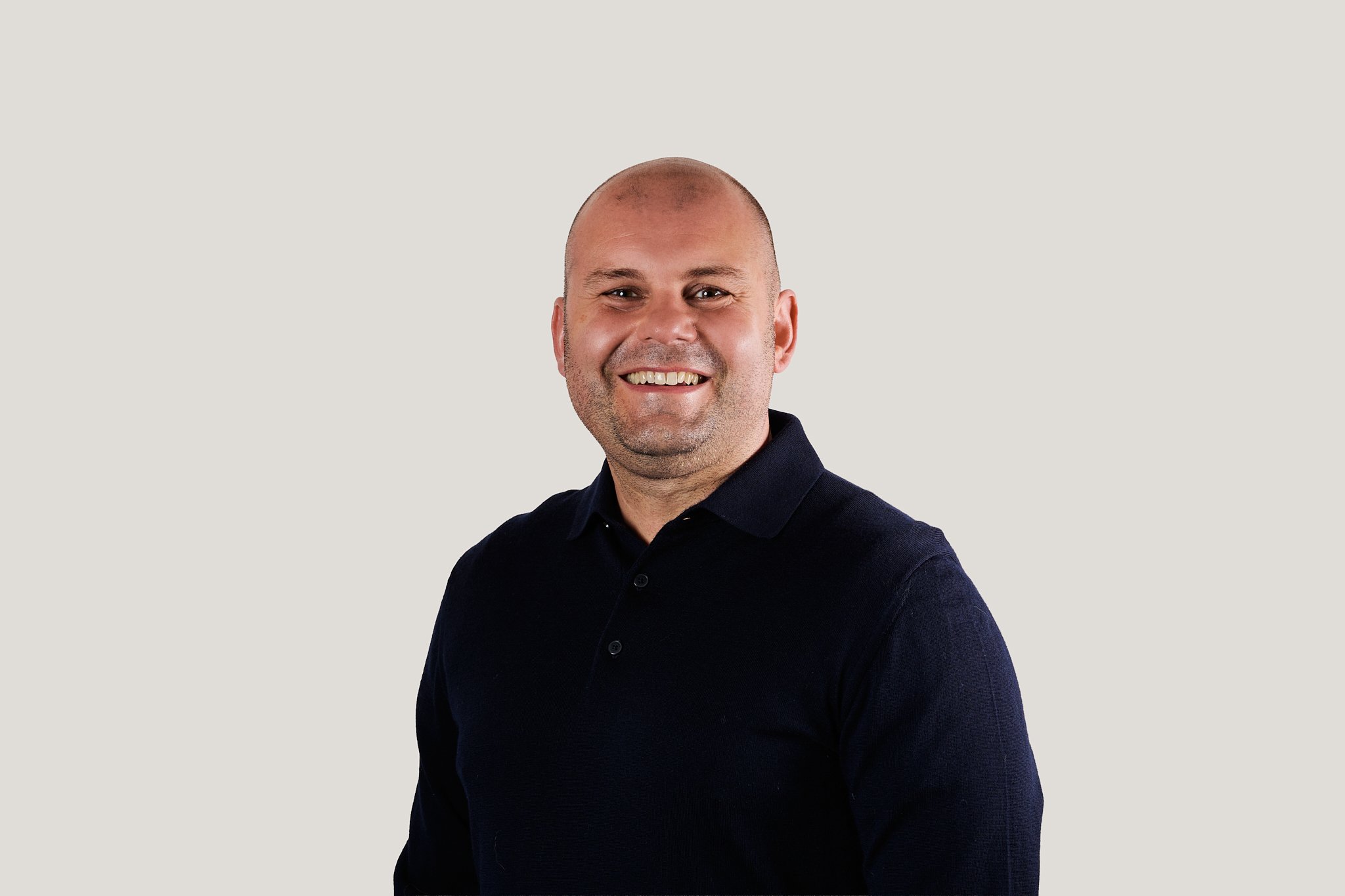 A smiling man with a bald head, wearing a dark collared shirt, standing against a plain light background.