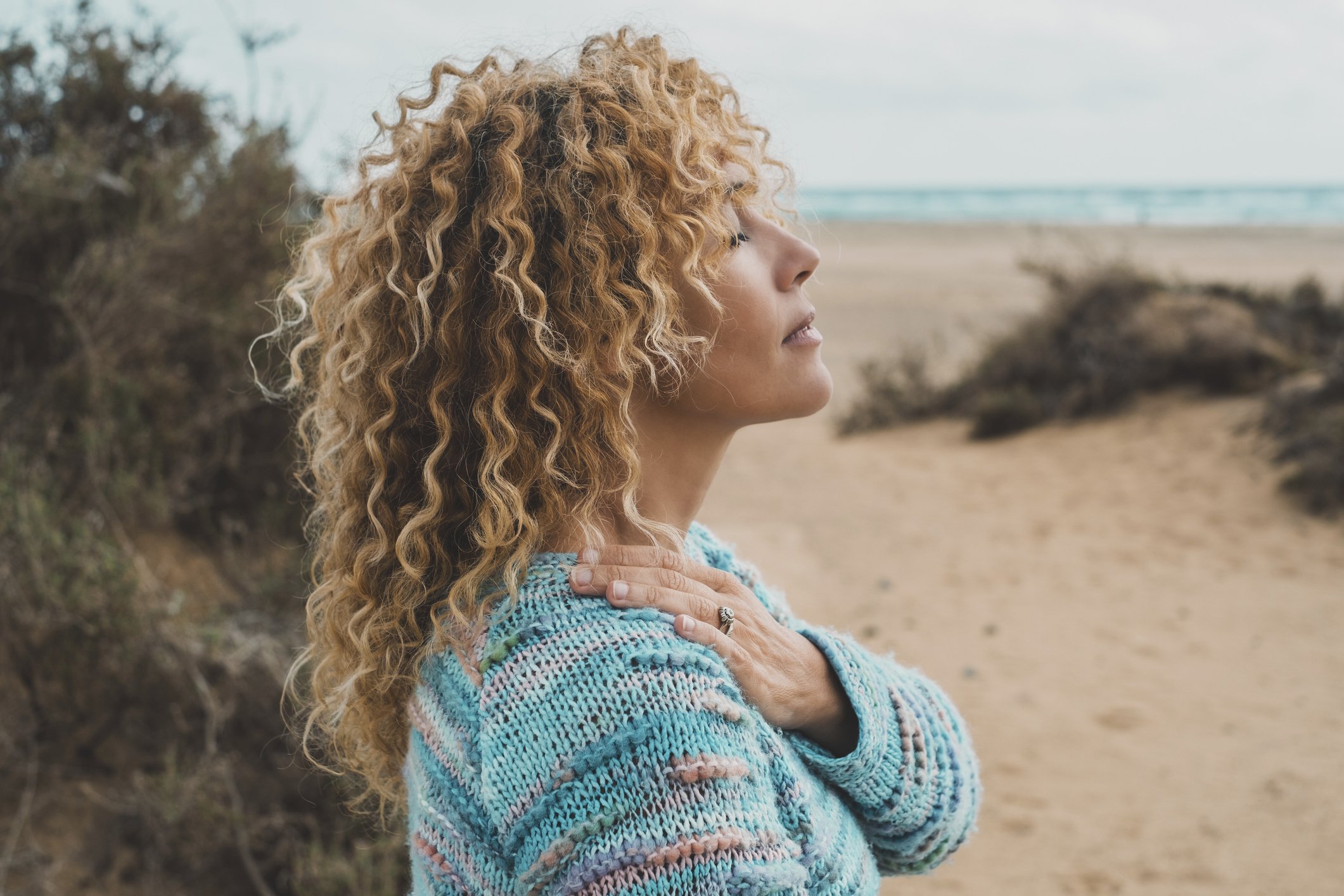 A woman with curly blonde hair standing on a beach, touching her shoulder, with the ocean in the background. Reflecting on her needs, finding space for herself, calm and peace after therapy session.