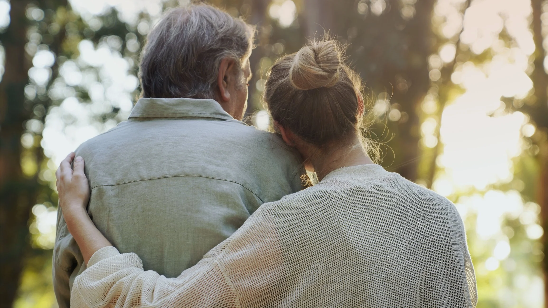 A woman and an elderly man sitting closely together outdoors, with the woman resting her head on the man's shoulder, surrounded by trees and sunlight filtering through. Considering their health journey, finding support coping with illness.