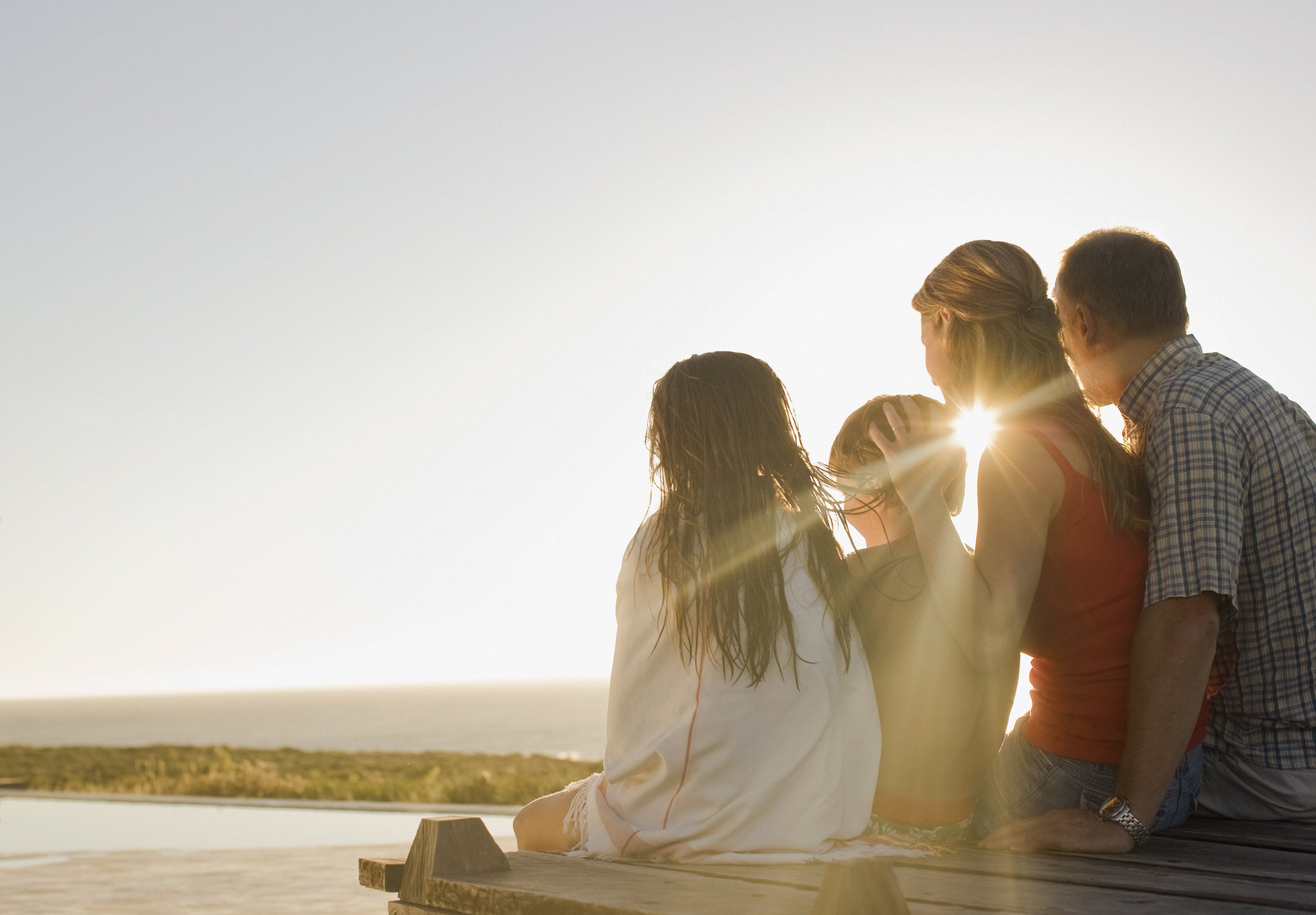 Family sitting on a dock near a swimming pool, watching the sunset, with the ocean in the background. Finding calm, engaged, happy together after family therapy.